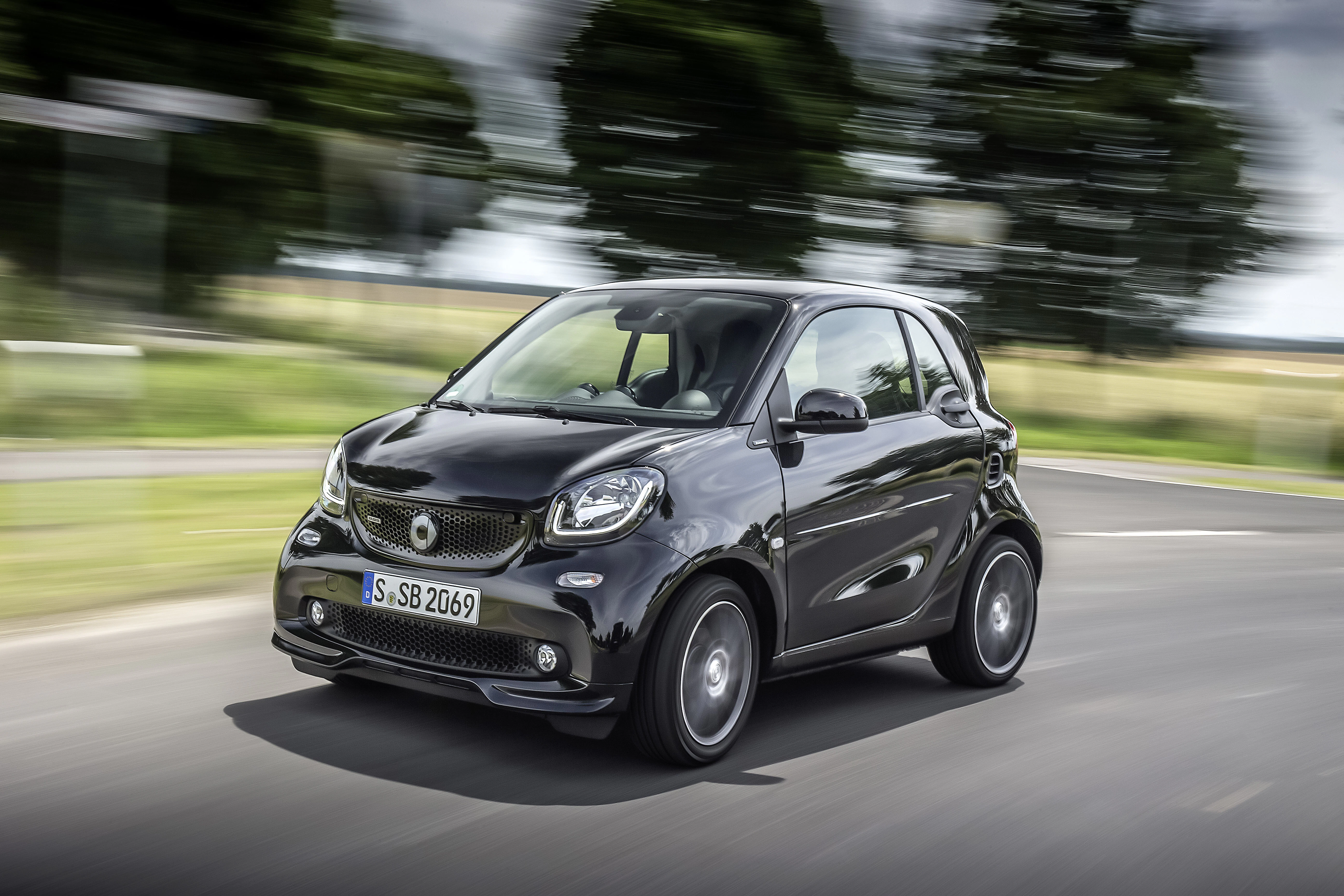 Schwarzer Smart Fortwo fährt auf einer Landstraße, unscharfer Hintergrund mit Bäumen und Wiesen. Dynamische Bewegung sichtbar.