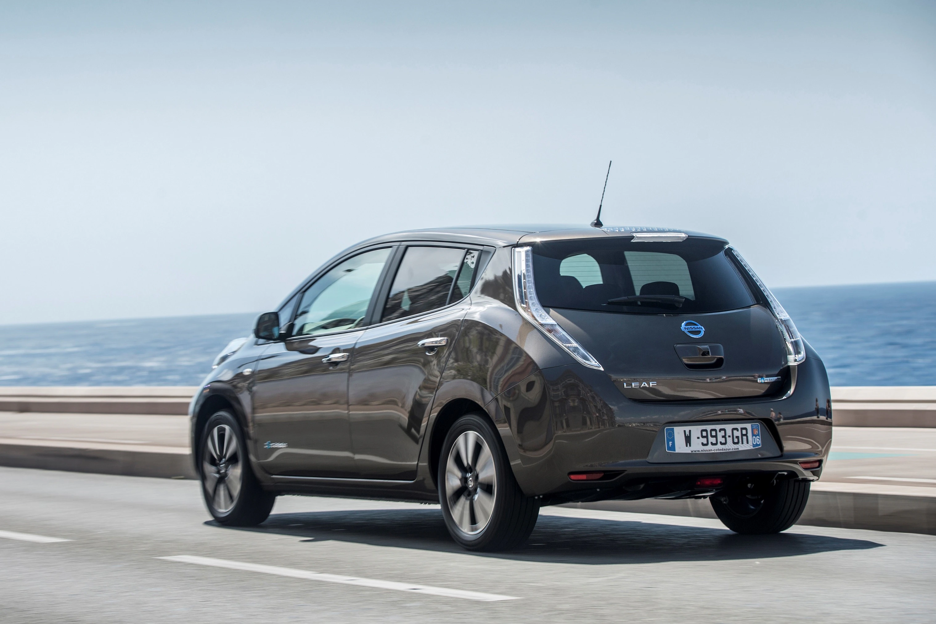 Ein brauner Nissan Leaf fährt auf einer Küstenstraße mit Blick auf das Meer unter klarem Himmel.