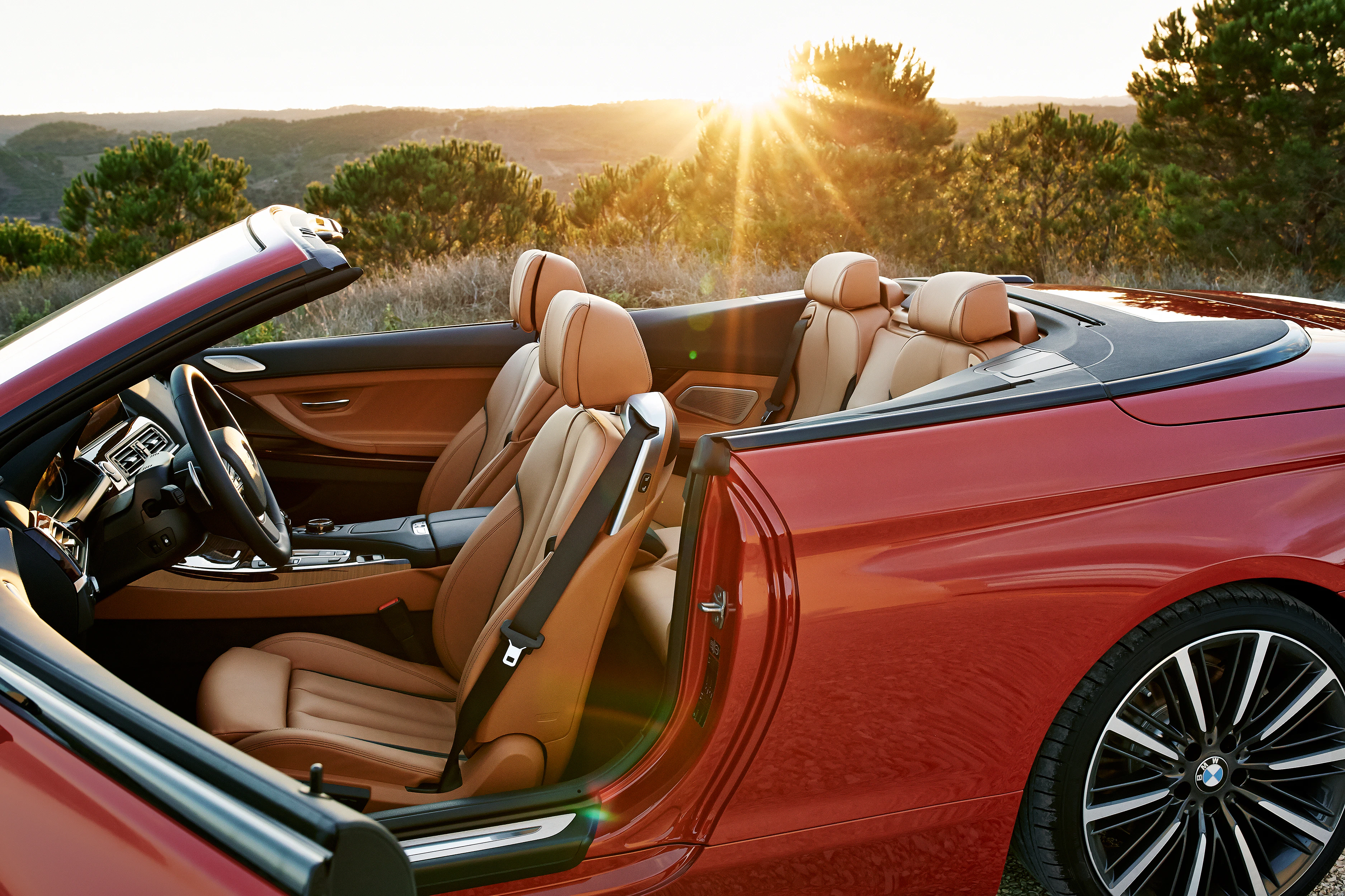 Rotes Cabrio mit beigen Ledersitzen, offen, bei Sonnenuntergang in einer Landschaft mit Bäumen und Hügeln im Hintergrund.