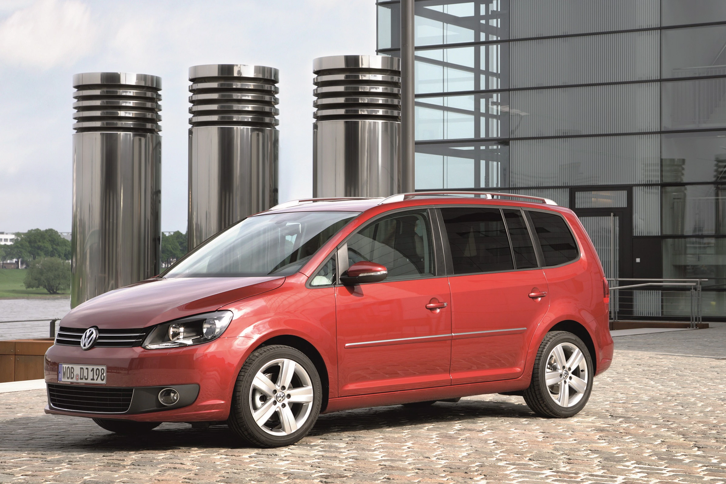 Roter VW Touran steht vor einem modernen Gebäude mit großen silbernen Lüftungstürmen auf gepflastertem Boden.