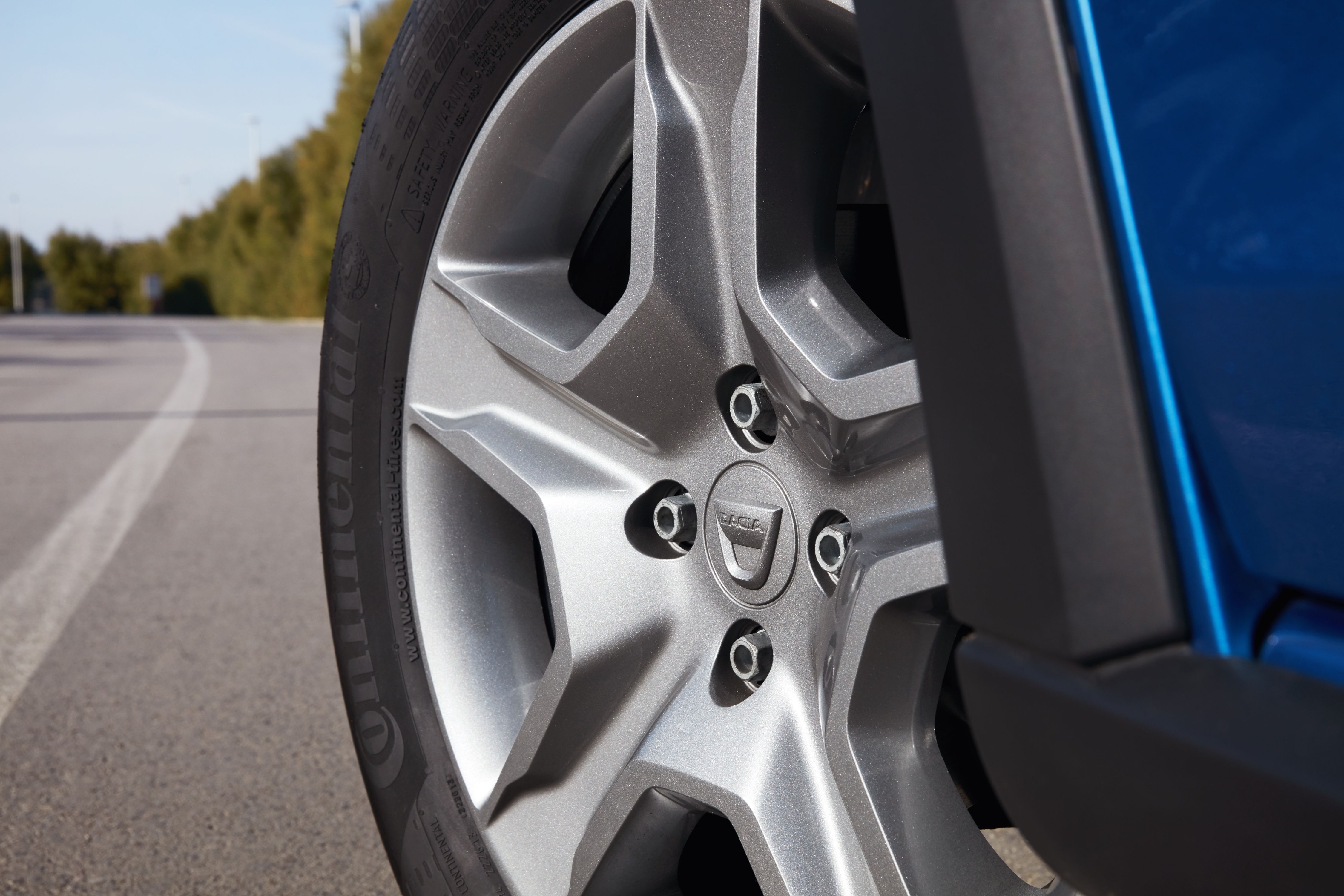Nahaufnahme eines silbernen Autoreifens mit einem stilisierten Radkasten. Im Hintergrund ist eine Straße und Vegetation zu sehen.