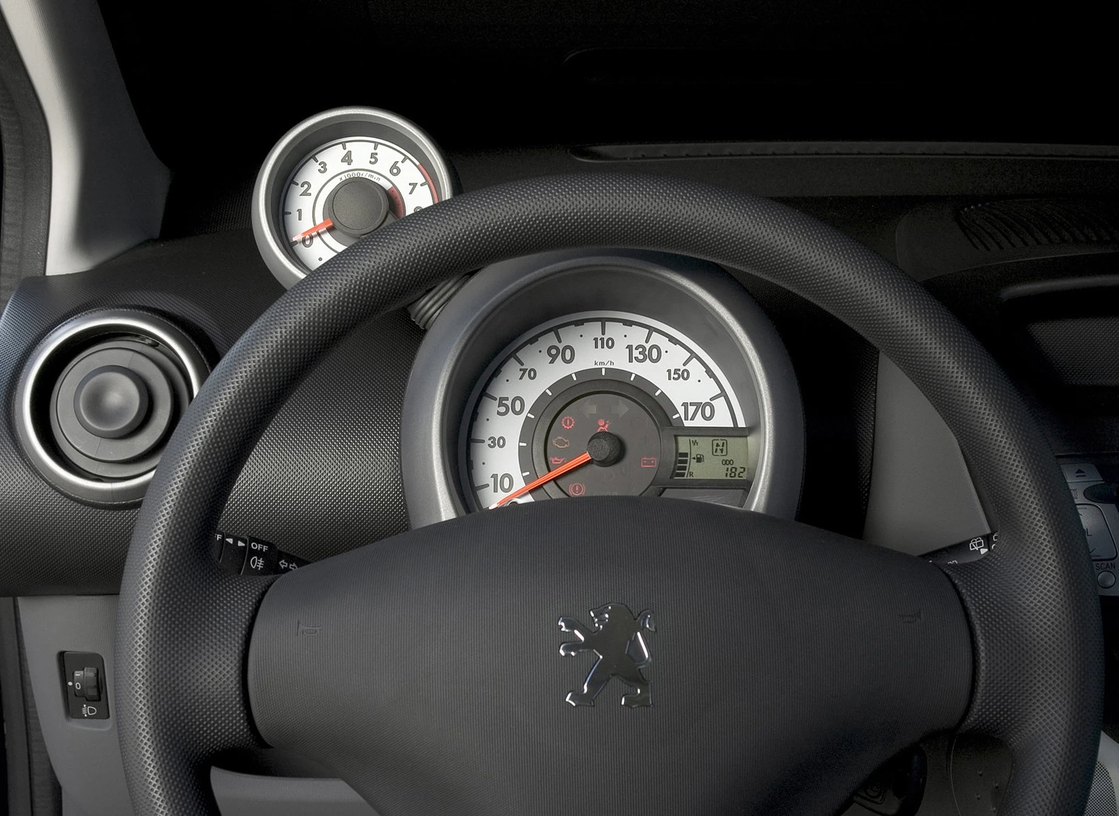 Lenkrad eines Autos mit Peugeot-Logo, Tachometer und Drehzahlmesser im Hintergrund, schwarz-graues Interieur.