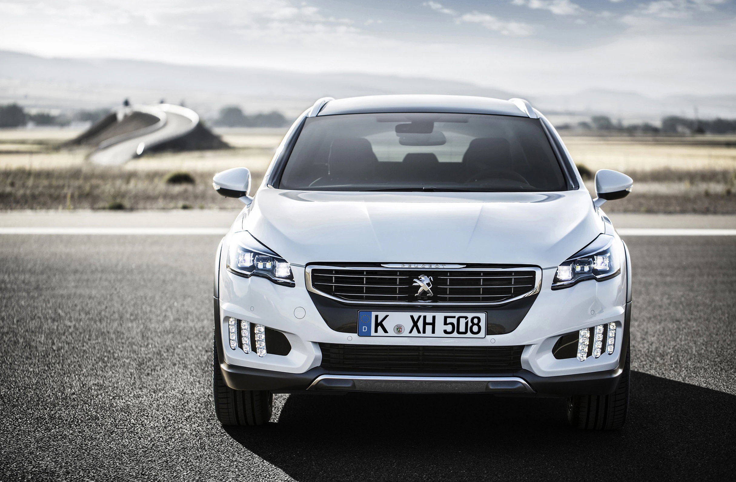 Ein weißer Peugeot steht auf einer Straße, bei sonnigem Wetter, vor einem leicht bewölkten Himmel und einer hügeligen Landschaft im Hintergrund.
