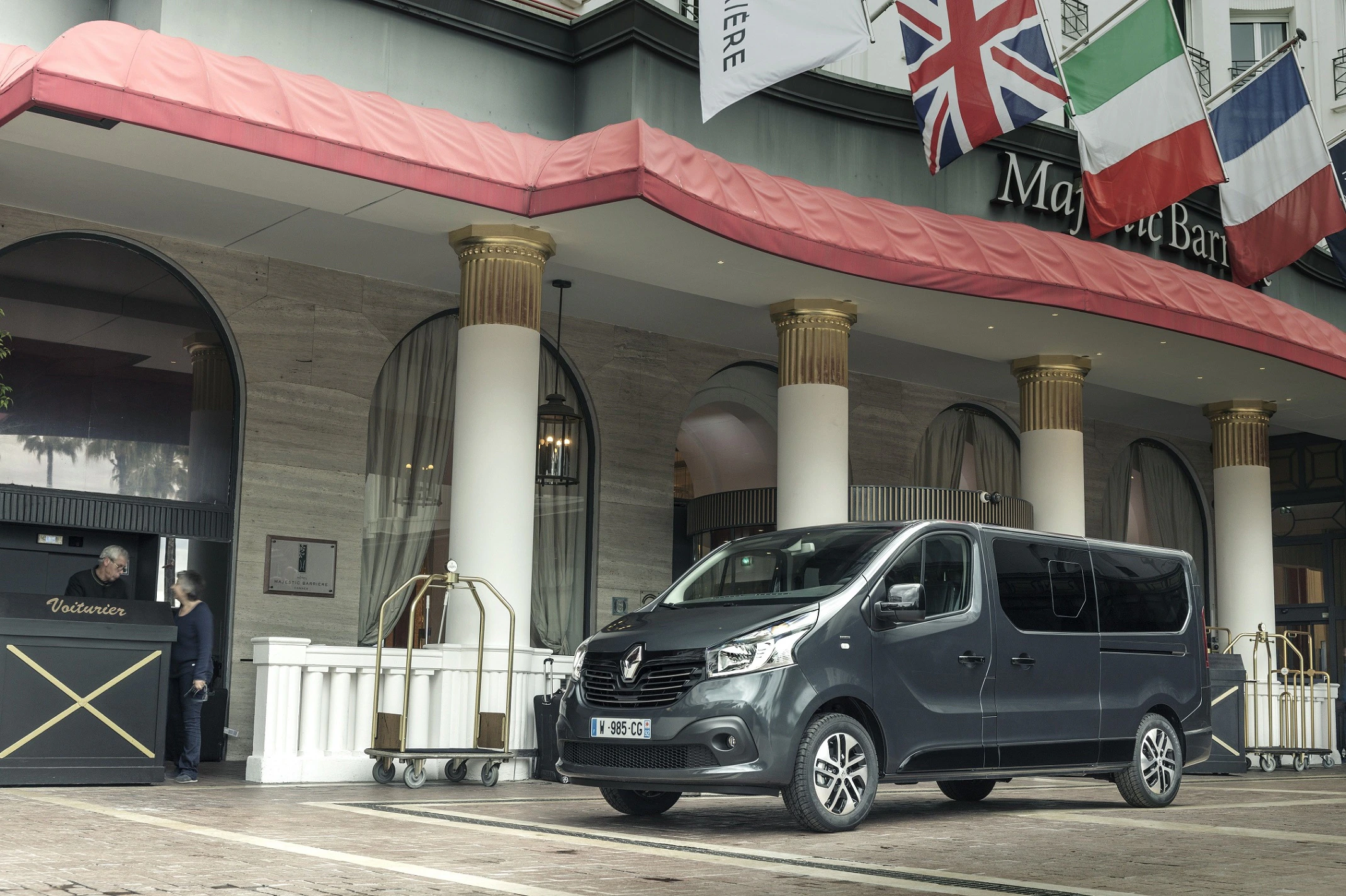 Ein dunkler Van steht vor einem eleganten Hotel mit mehreren Flaggen und Säulen. Im Hintergrund sind ein Portier und Gepäckwagen zu sehen.