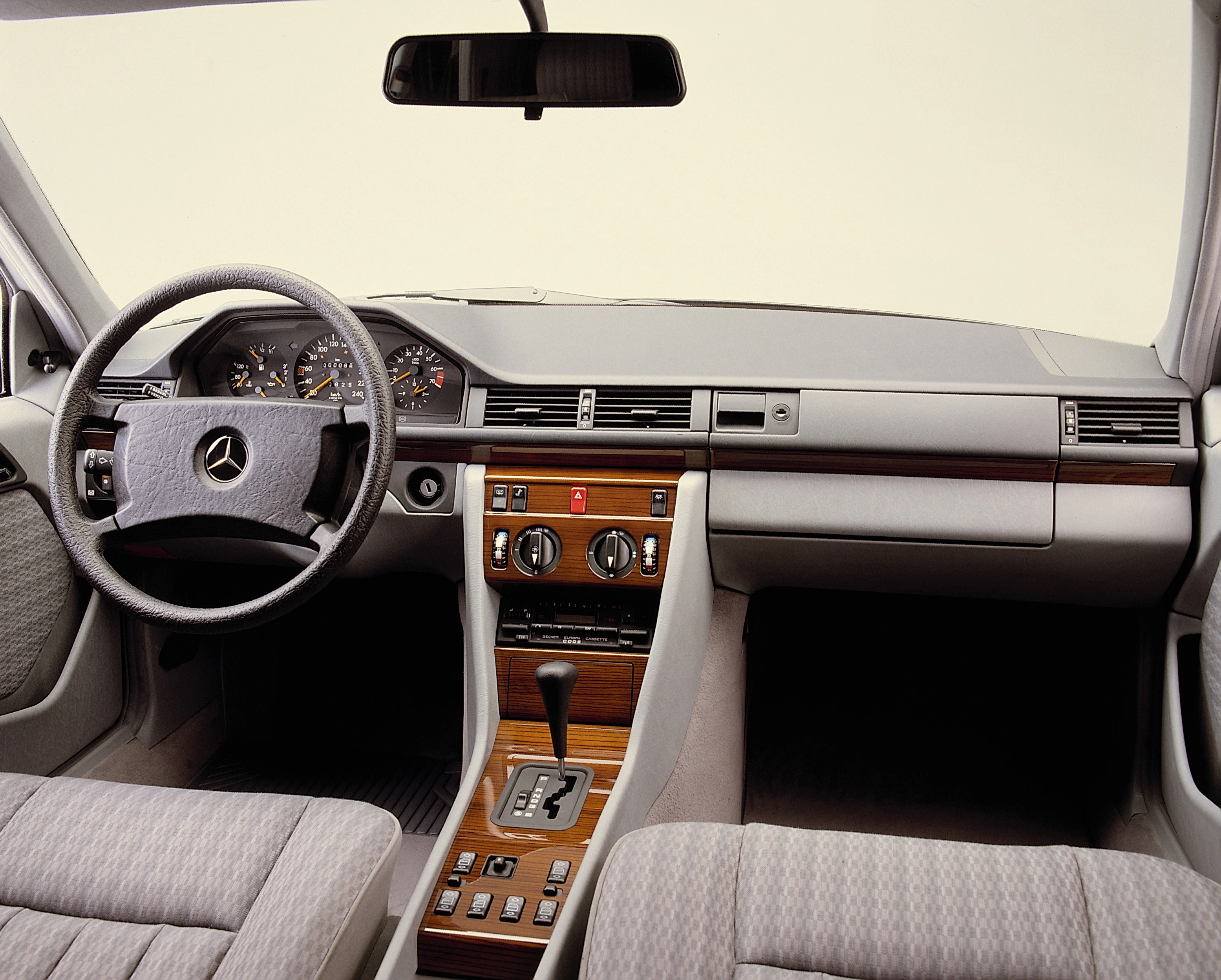 Innenraum eines Autos mit Mercedes-Logo auf dem Lenkrad, Holzverkleidung auf der Mittelkonsole, analogen Armaturen und hellen Stoffpolstern.