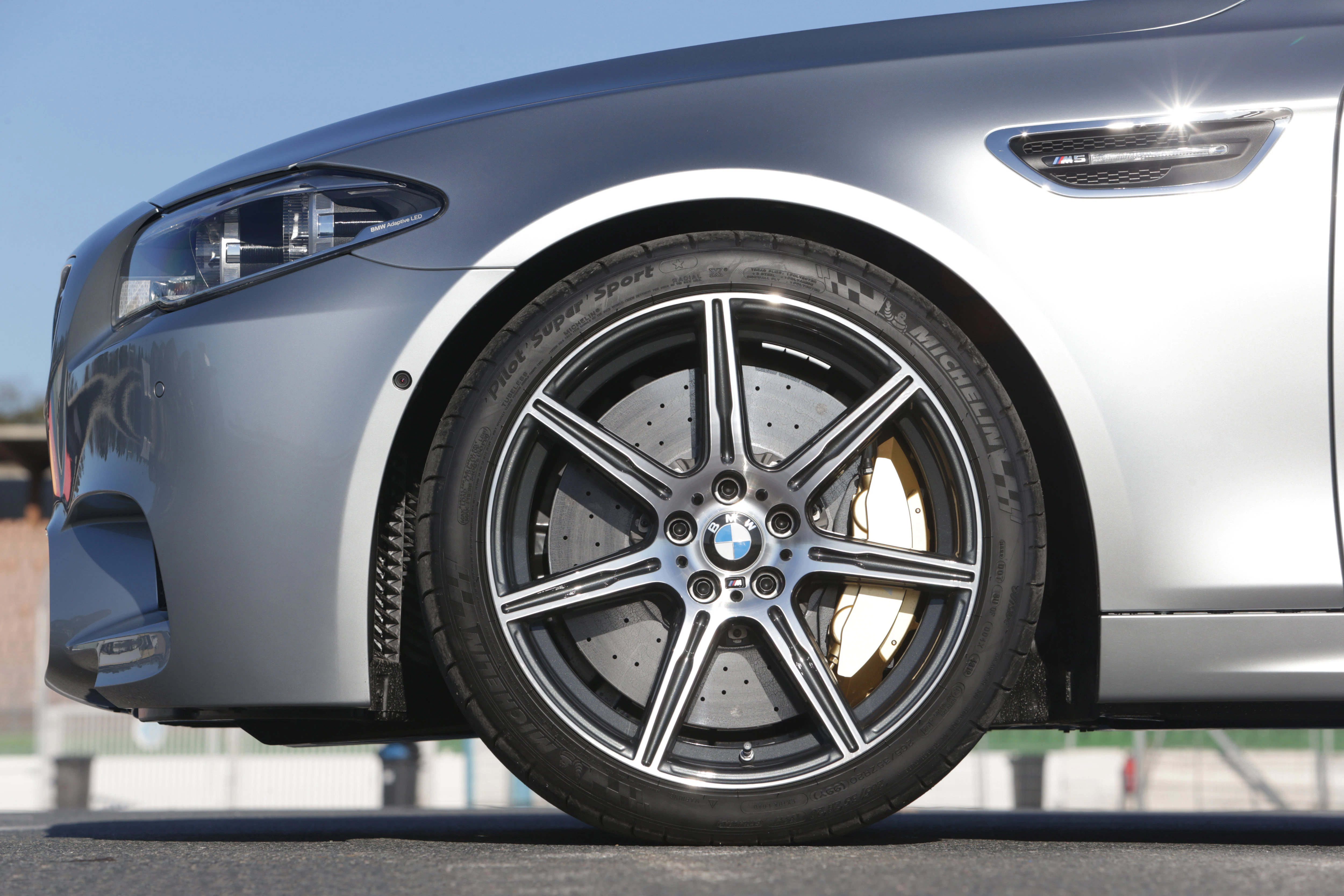 Nahaufnahme eines silbernen BMWs mit markanten Alufelgen und sichtbaren Bremssätteln, auf einer Straße bei blauem Himmel.