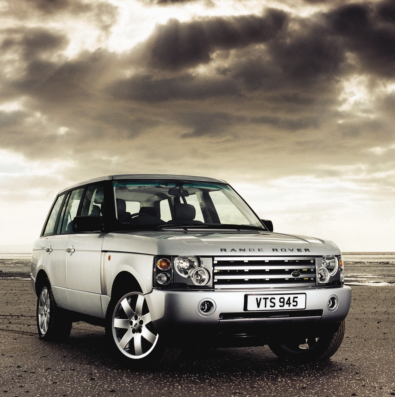 Ein silberner Range Rover steht vor einem dramatisch bewölkten Himmel auf einem Strand. Das Auto hat ein britisches Kennzeichen VTS 945.