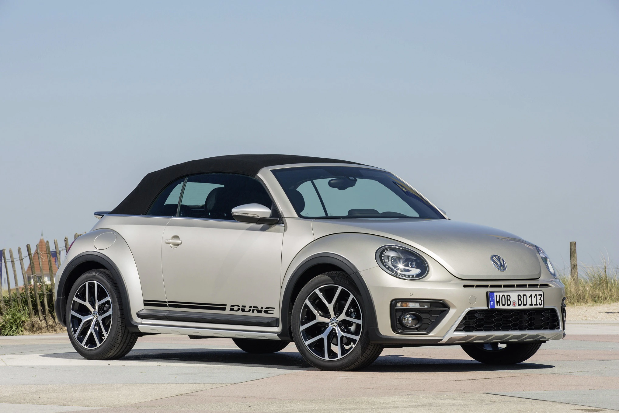 Silberner VW Beetle Cabrio parkt auf einem sonnigen Platz. Der Himmel ist klar, und im Hintergrund ist ein Zaun erkennbar.