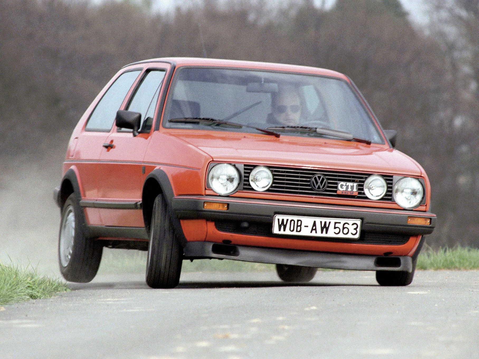 Roter VW Golf GTI der 1980er Jahre, fährt dynamisch über eine Landstraße. Der Himmel ist bewölkt und Bäume im Hintergrund.