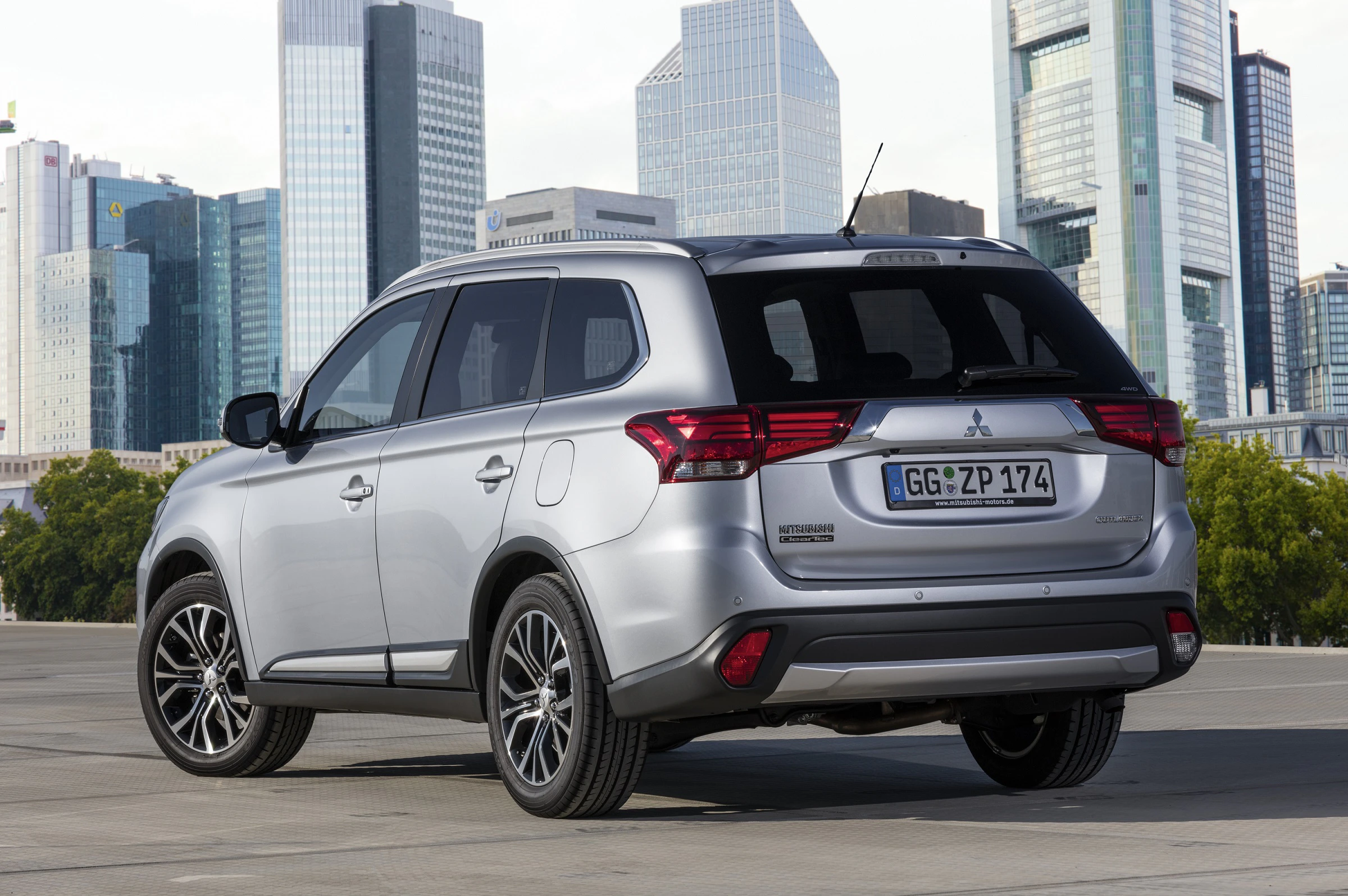 Silberner SUV Mitsubishi Outlander PHEV auf einem Parkplatz, im Hintergrund moderne Hochhäuser und blauer Himmel.