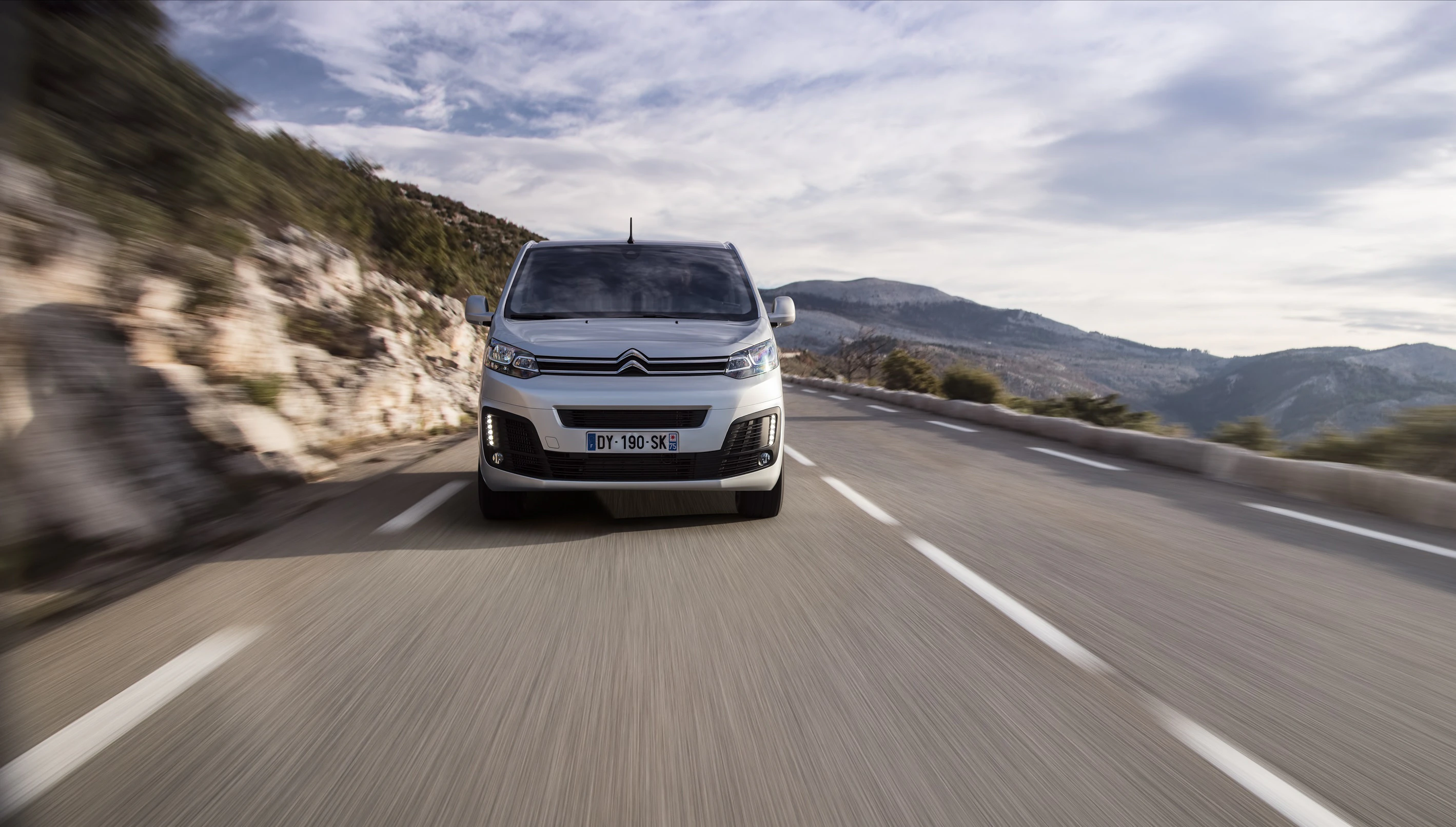 Silberner Citroën-Transporter fährt auf einer kurvigen Bergstraße mit felsigen Klippen und bewölktem Himmel im Hintergrund.