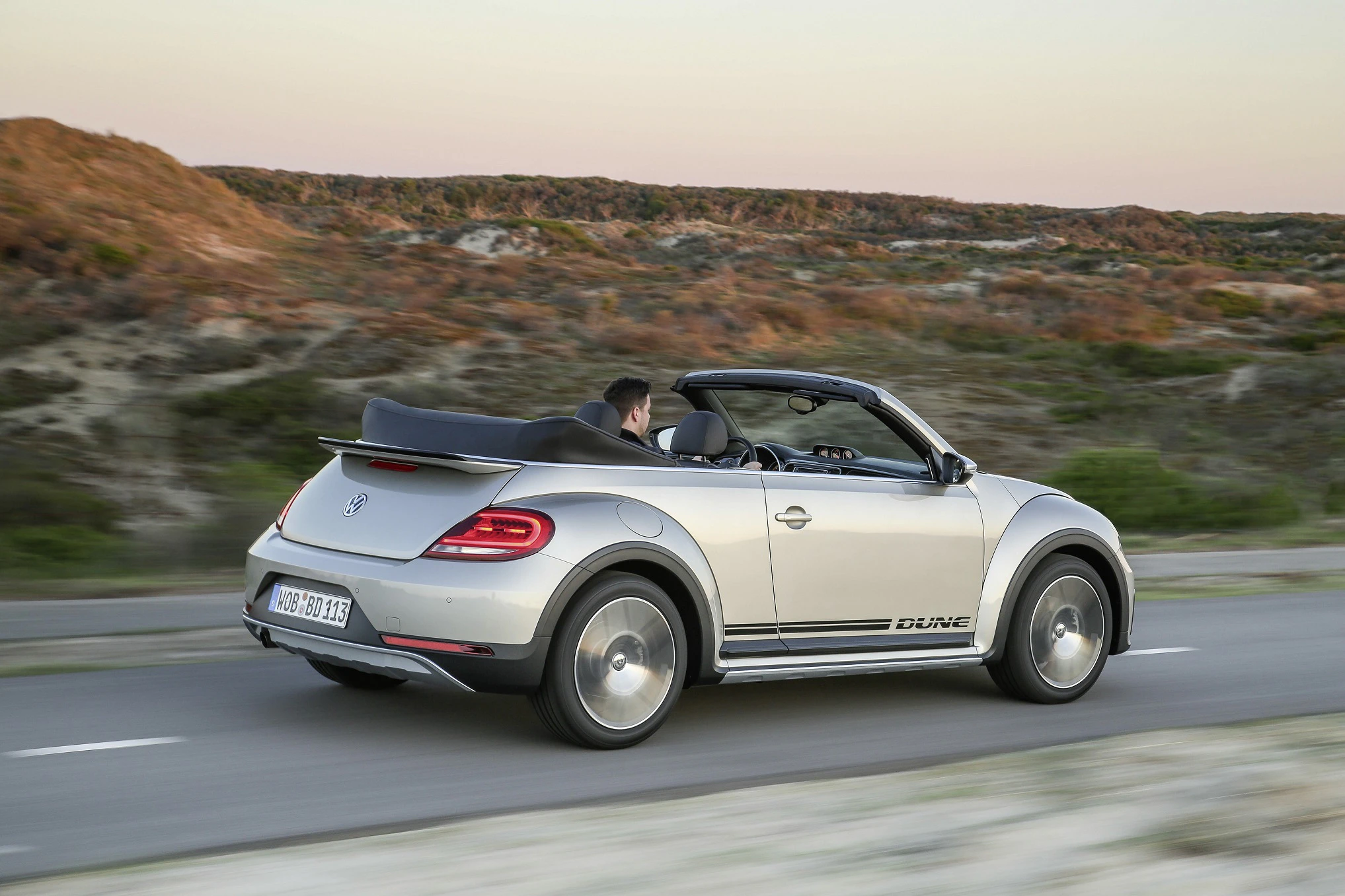 Silberner VW Beetle Cabrio fährt bei Sonnenuntergang auf einer Landstraße an einer hügeligen Landschaft vorbei.