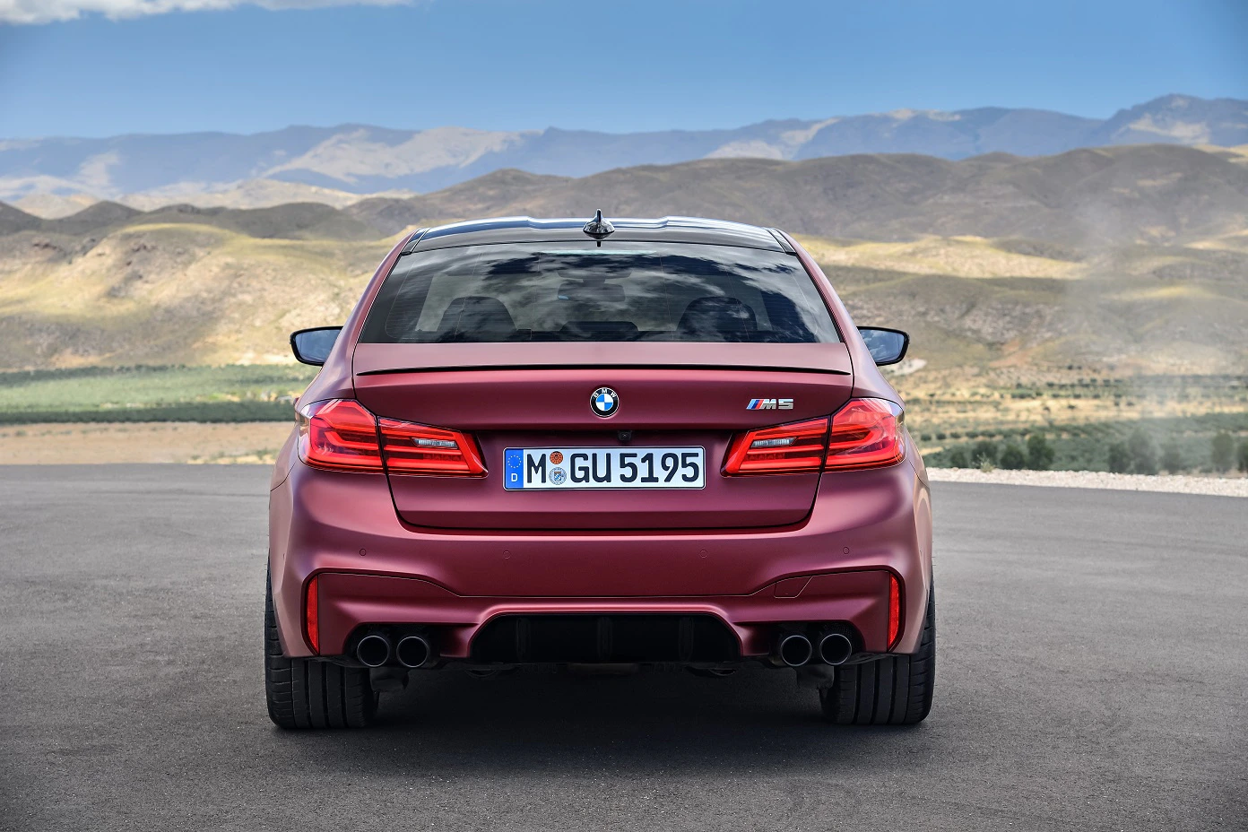 Roter BMW M5 steht auf einer Straße, mit Bergen und blauem Himmel im Hintergrund. Rückansicht des Autos zeigt sportliches Design und Auspuffanlagen.