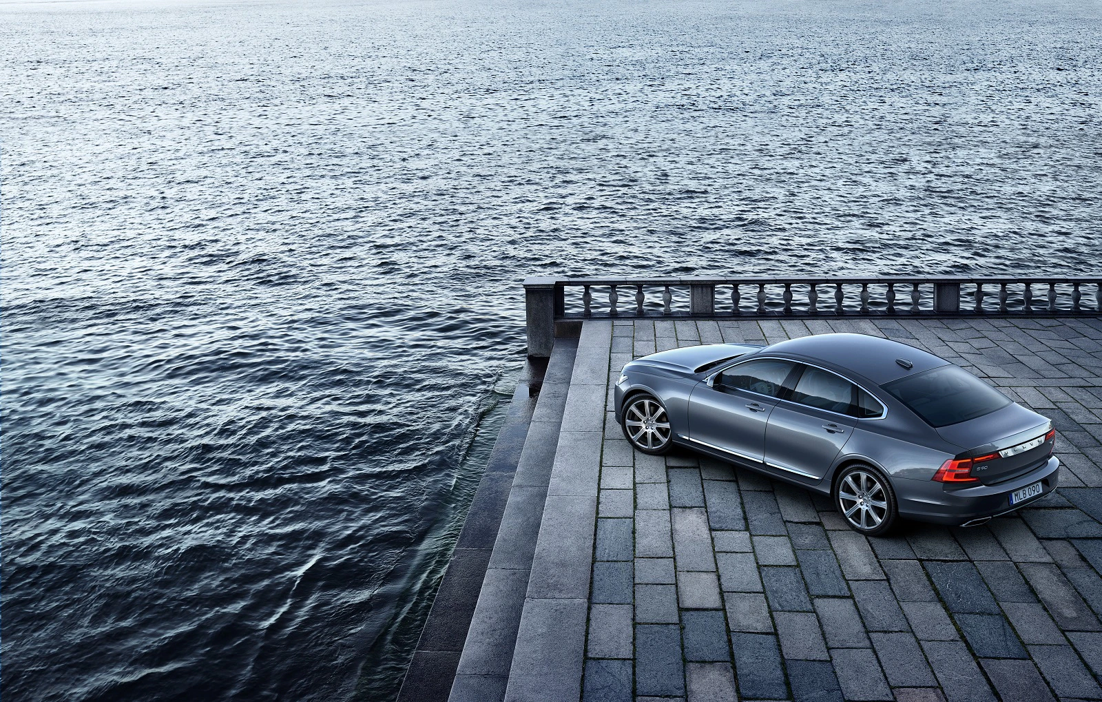 Silbernes Auto auf gepflastertem Platz direkt am Wasser, mit weitläufigem Meer und bewölktem Himmel im Hintergrund.