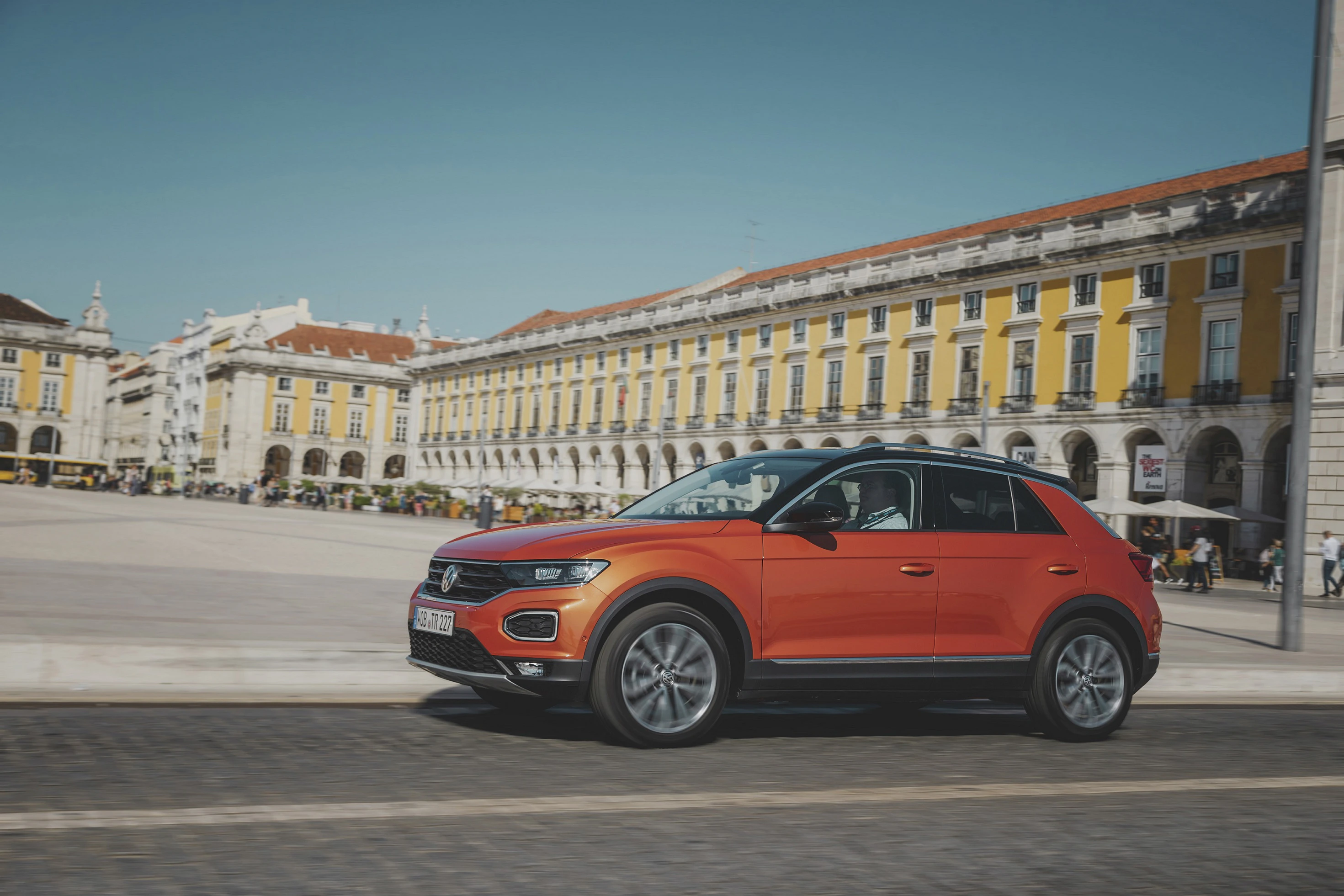 Ein oranger SUV fährt durch einen sonnigen Platz mit historischen Gebäuden im Hintergrund.