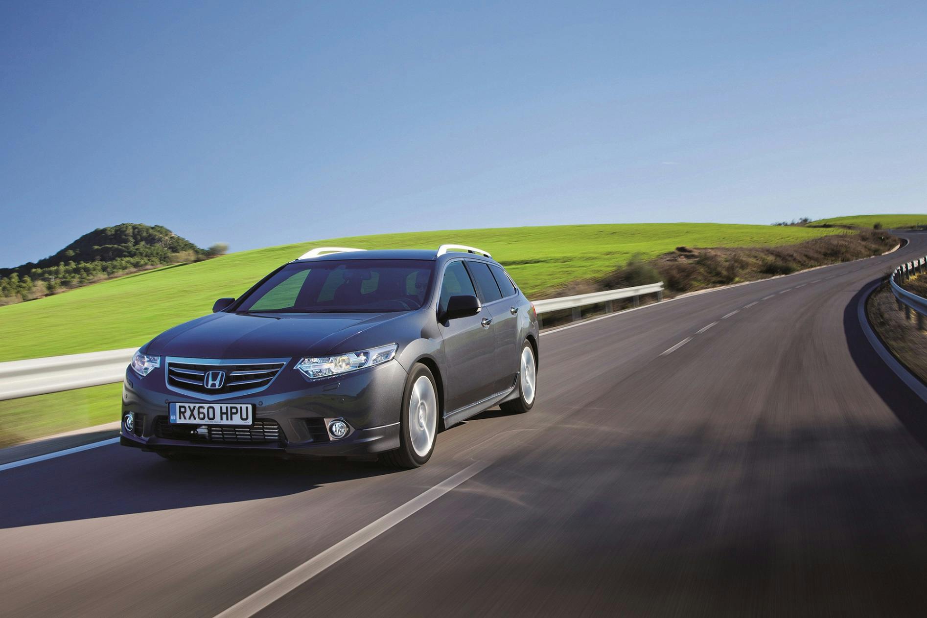 Honda-Kombi fährt auf einer Landstraße durch eine grüne, hügelige Landschaft unter klarem, blauem Himmel.