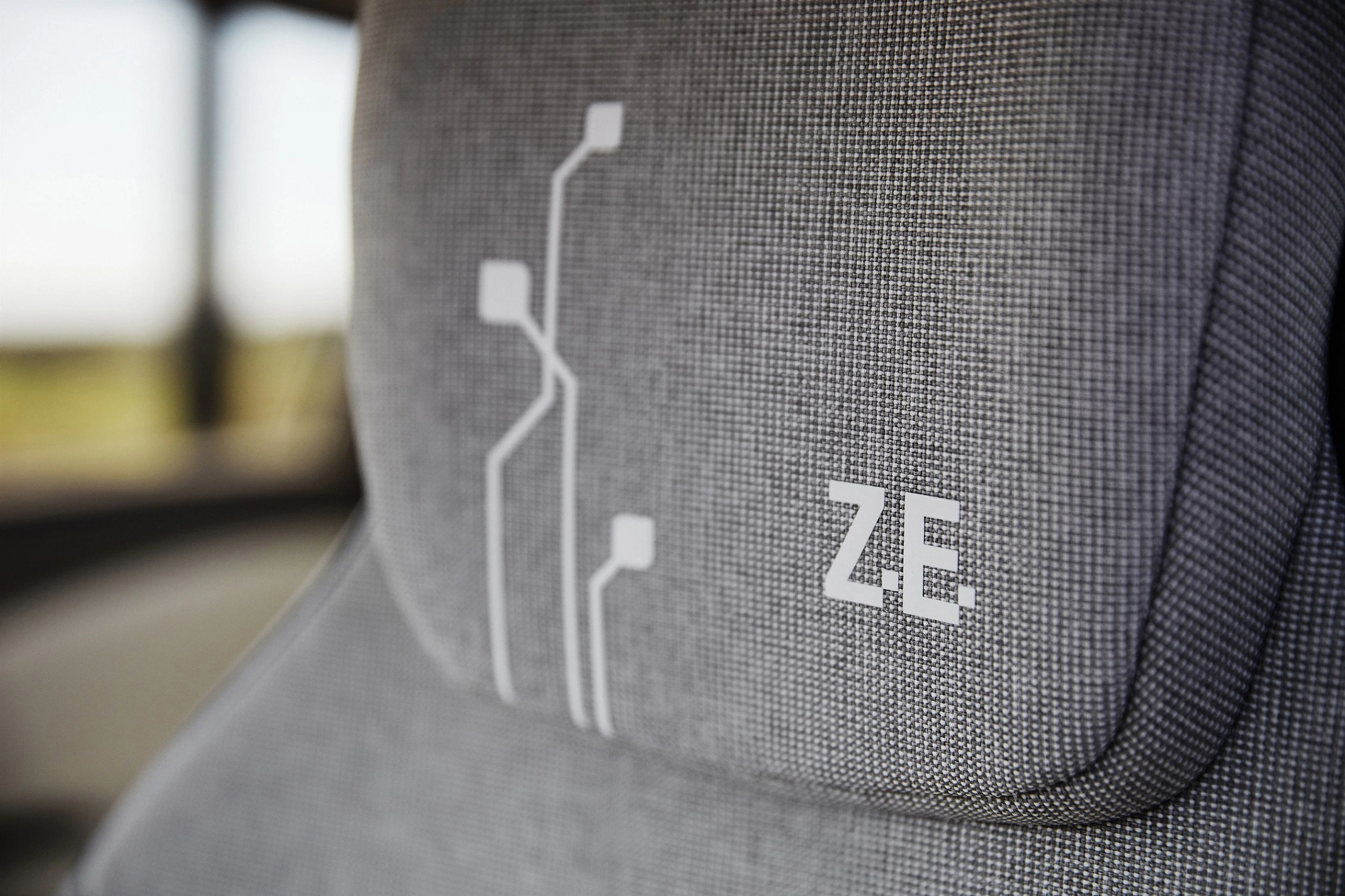 Grauer Autositz mit weißem grafischem Muster und den Buchstaben "ZE" darauf. Hintergrund zeigt verschwommenes Licht.