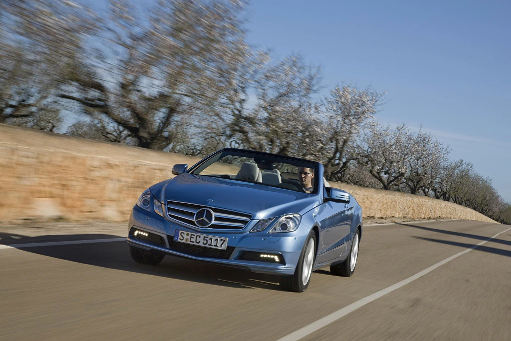 Blaues Mercedes-Cabriolet fährt auf einer Landstraße unter blauem Himmel, im Hintergrund blühende Bäume und Steinmauer.