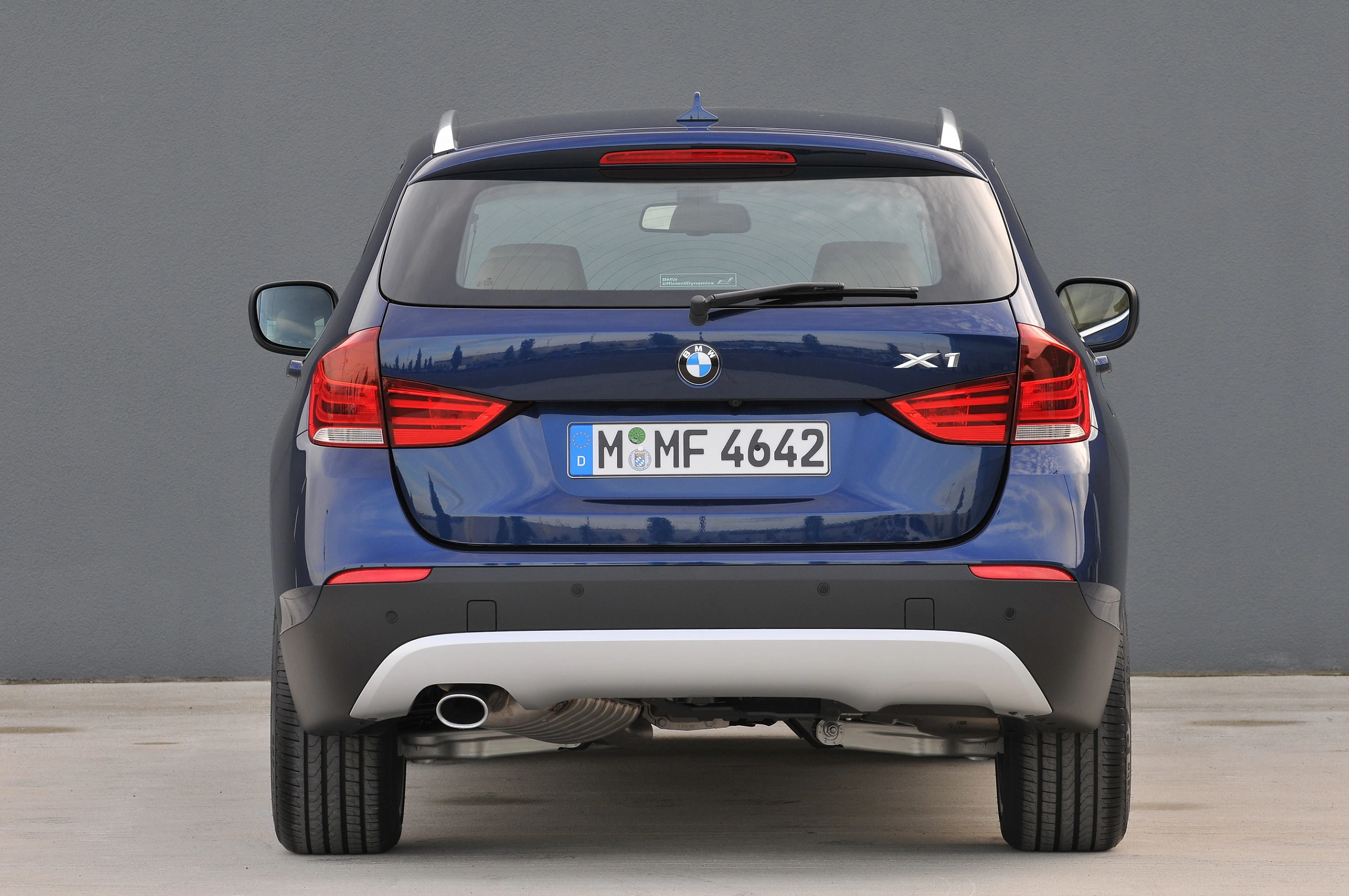 Blauer BMW X1 von hinten, geparkt vor einer grauen Wand. Ansicht zeigt Rückleuchten, Auspuff und Nummernschild.