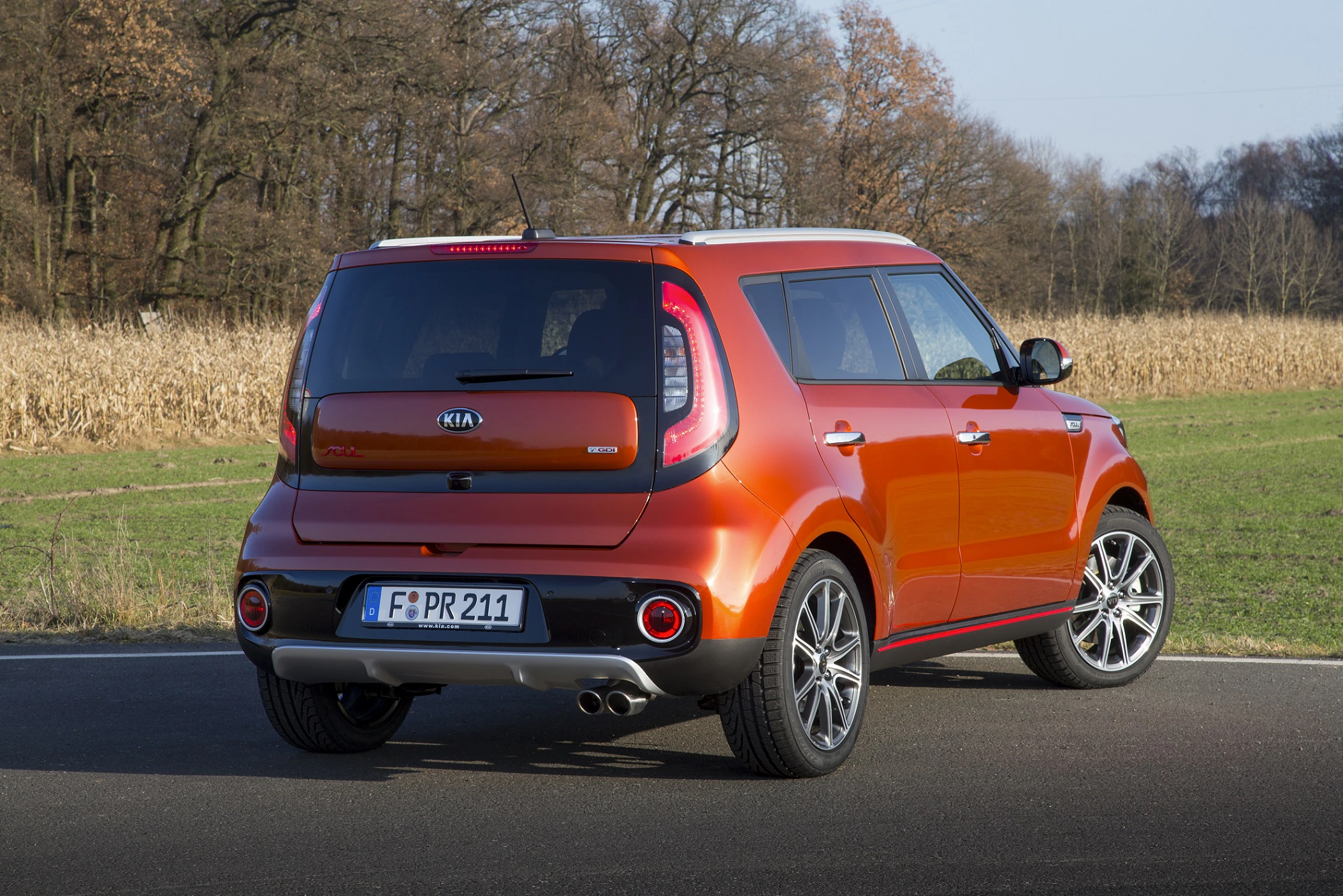 Ein orangefarbener Kia Soul steht auf einer Straße, umgeben von Feldern und Bäumen im Hintergrund.