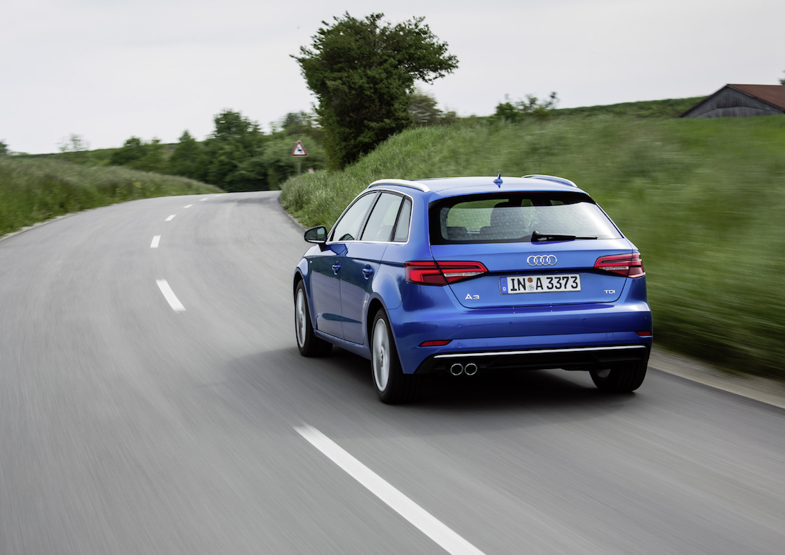 Blauer Audi auf einer kurvigen Landstraße, umgeben von grüner Landschaft und bewölktem Himmel. Das Auto fährt nach links.