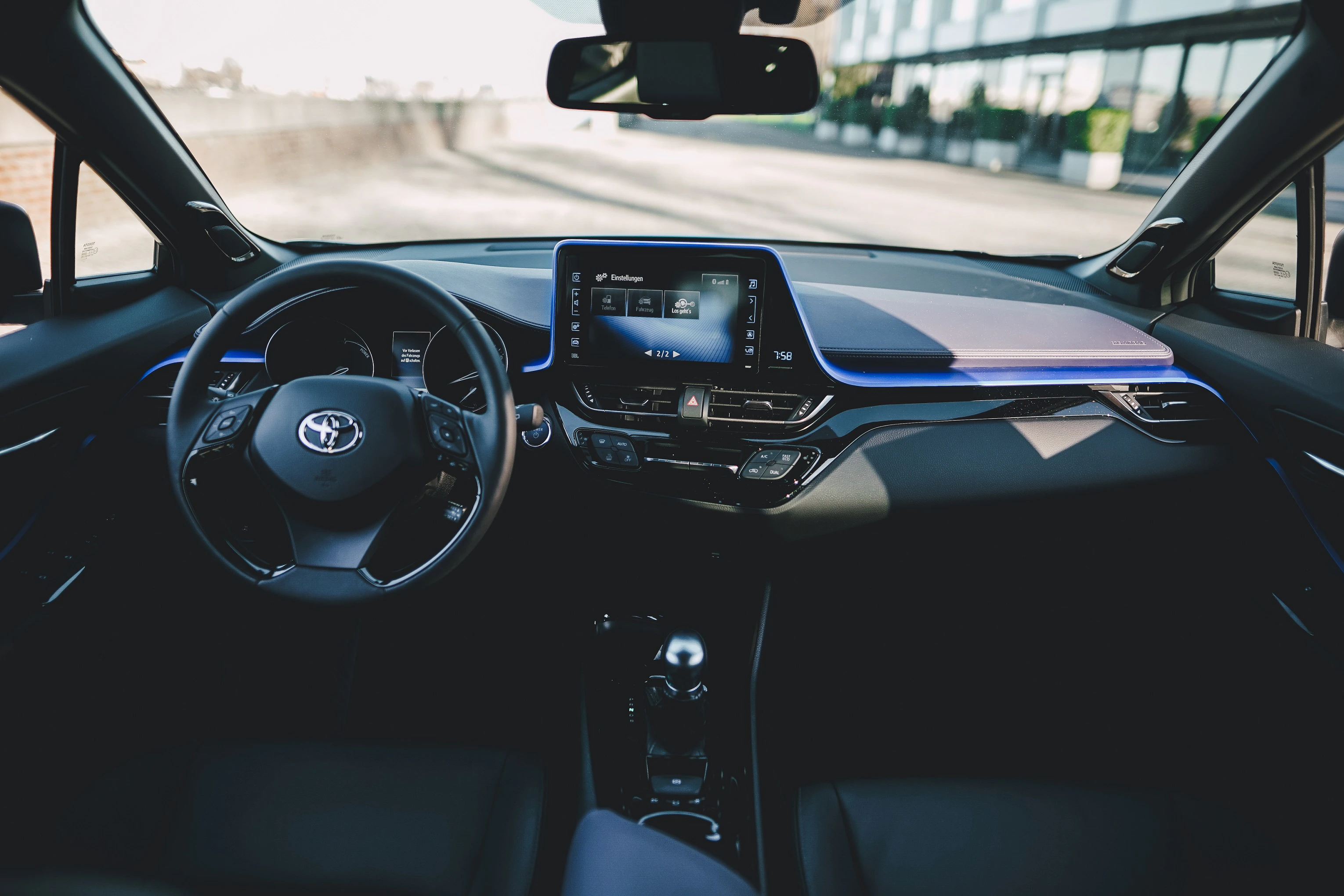 Innenraum eines modernen Autos mit Toyota-Logo. Digitales Display in der Mitte, Lenkrad links. Blaue Akzente auf dem Armaturenbrett.