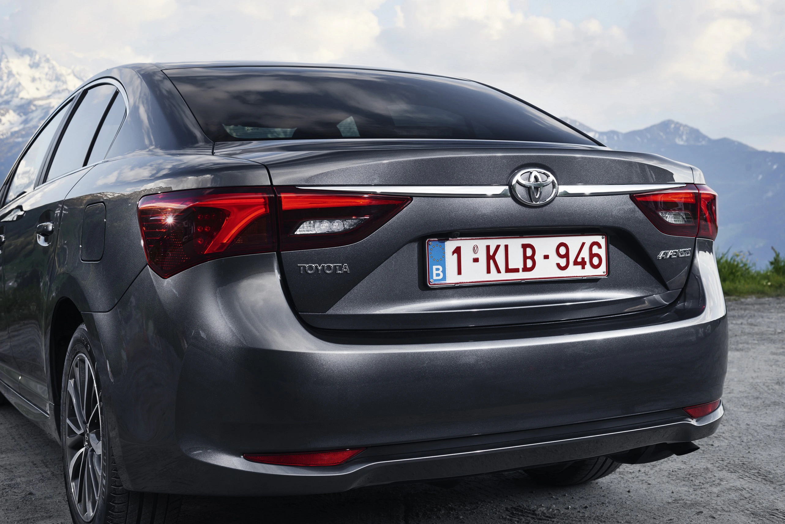 Graues Auto der Marke Toyota mit belgischem Kennzeichen, geparkt vor einer Berglandschaft unter bewölktem Himmel.