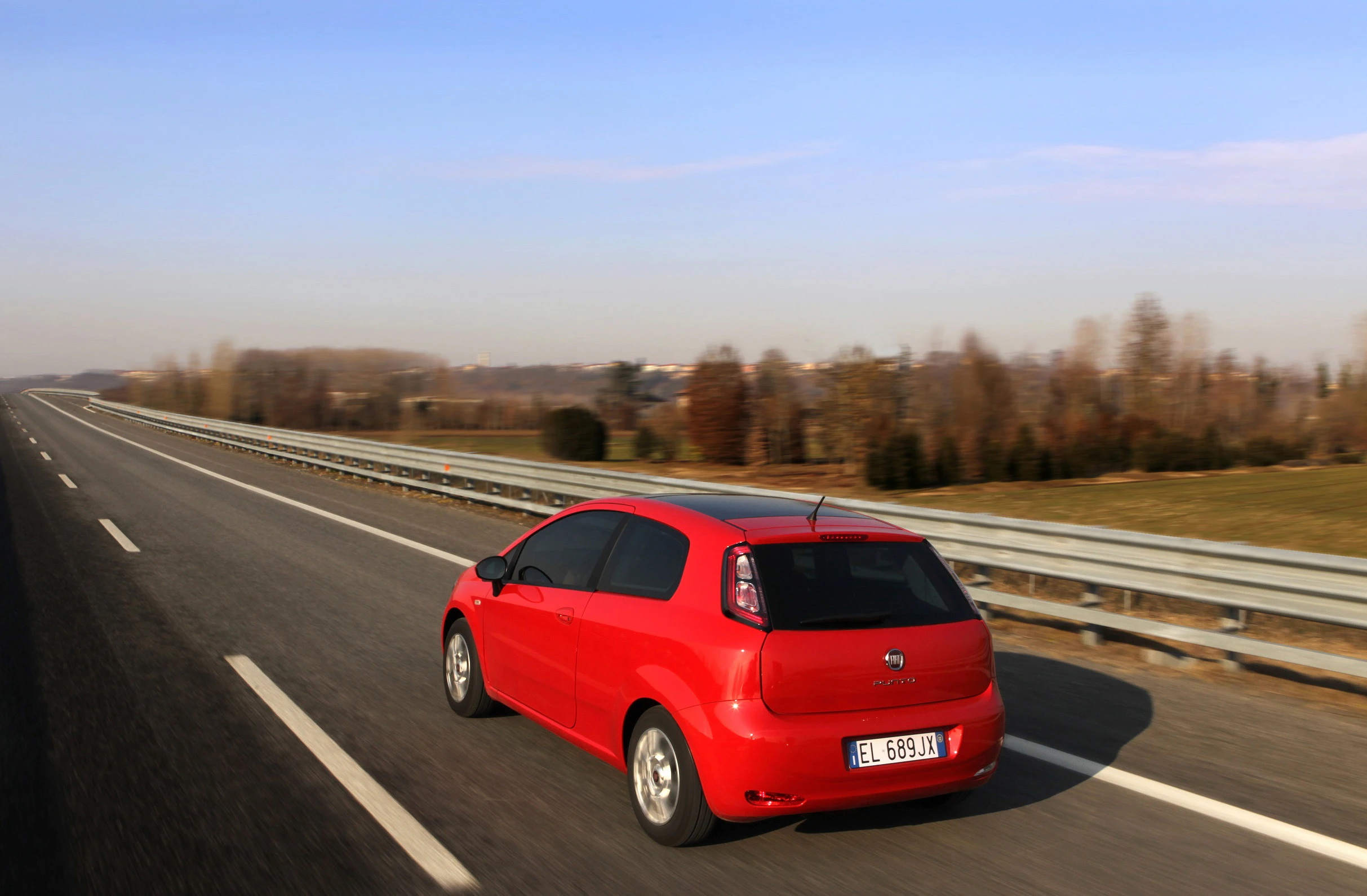 Rotes Auto fährt auf zweispuriger Autobahn durch ländliche Landschaft unter blauem Himmel.