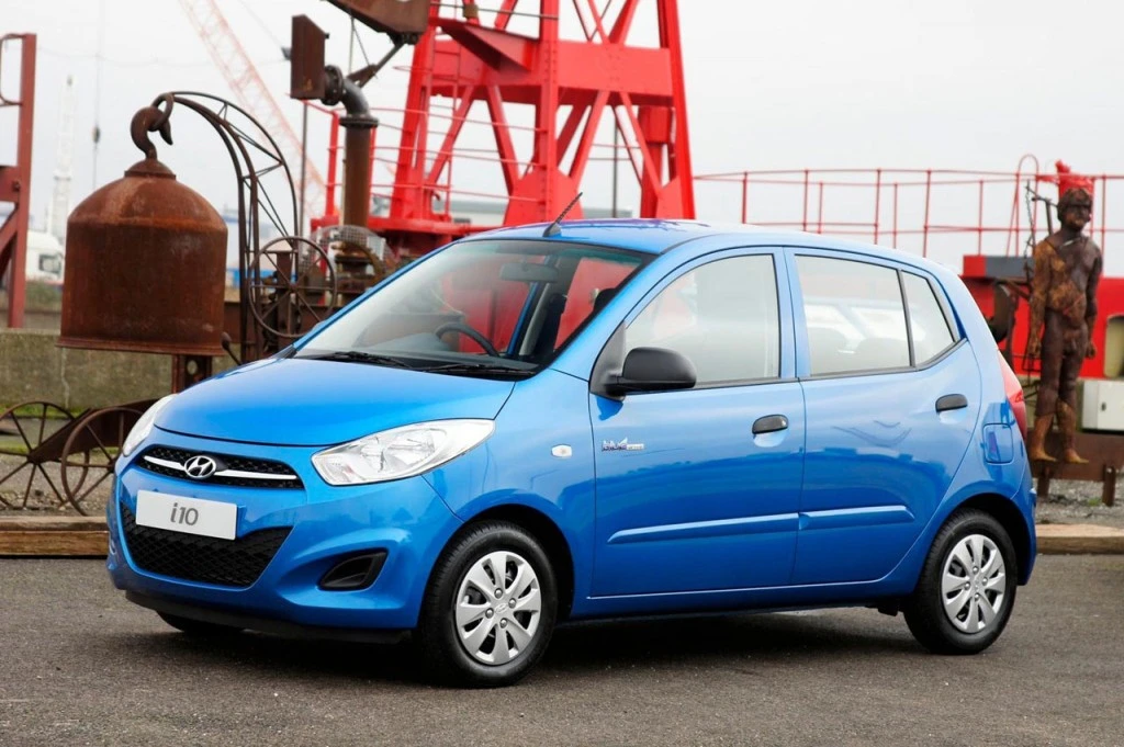 Blauer Hyundai i10 steht auf einem Parkplatz vor industrieller Kulisse mit roten Stahlstrukturen im Hintergrund.