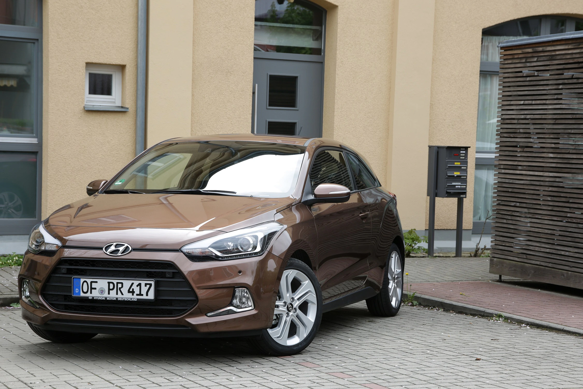Brauner Hyundai i20 parkt vor einem Gebäude mit gelber Fassade und Holzelementen, deutsches Kennzeichen sichtbar.
