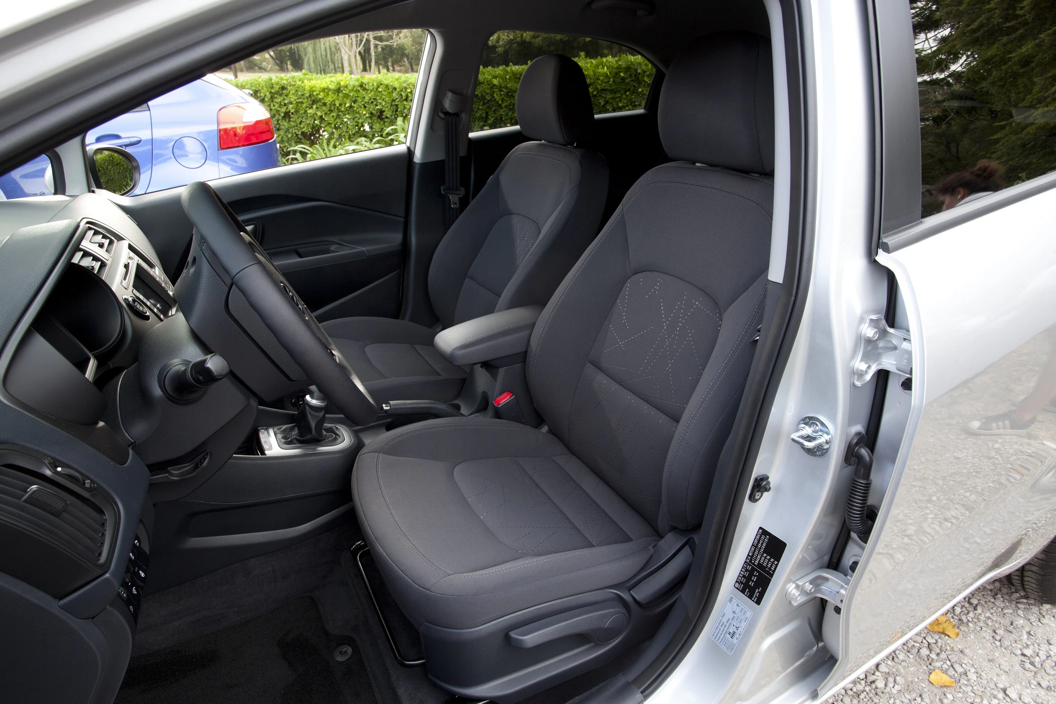Innenraum eines silbernen Autos mit geöffneten Türen, grauen Sitzen und einem dunklen Armaturenbrett. Ein blaues Auto ist im Hintergrund sichtbar.