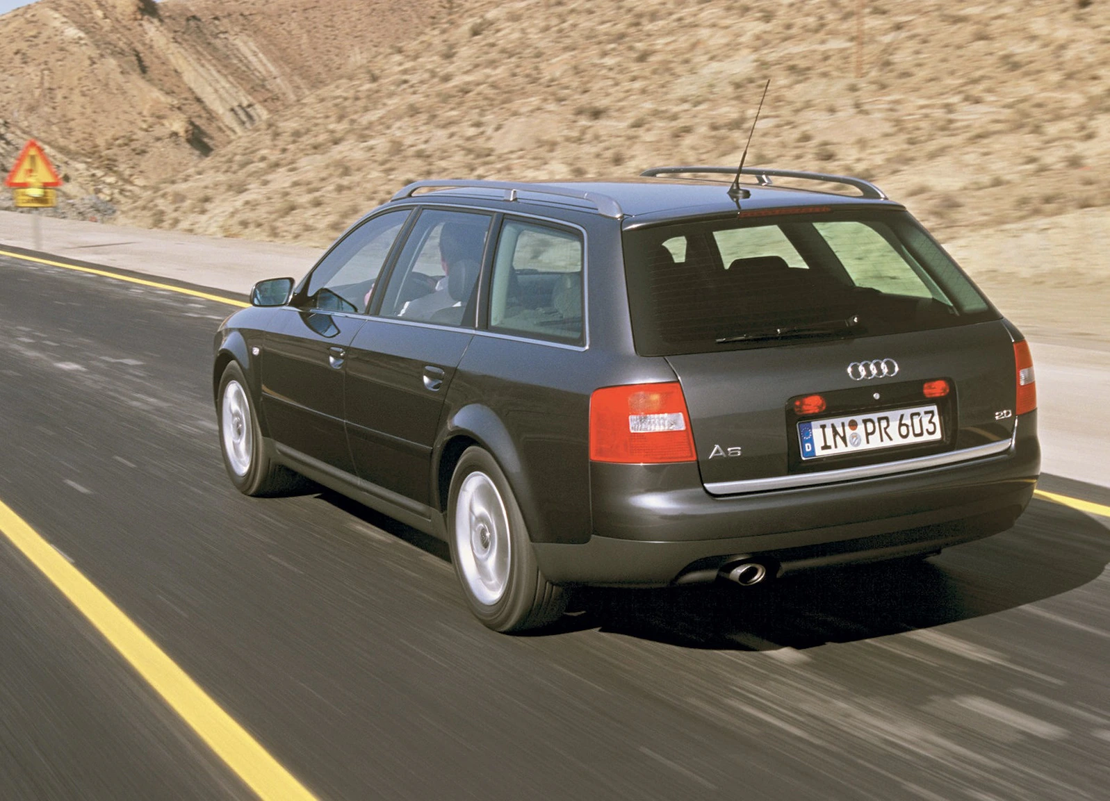 Audi A6 Kombi fährt auf einer Straße durch eine wüstenartige Landschaft, bergauf in Richtung eines Warnschilds am Straßenrand.