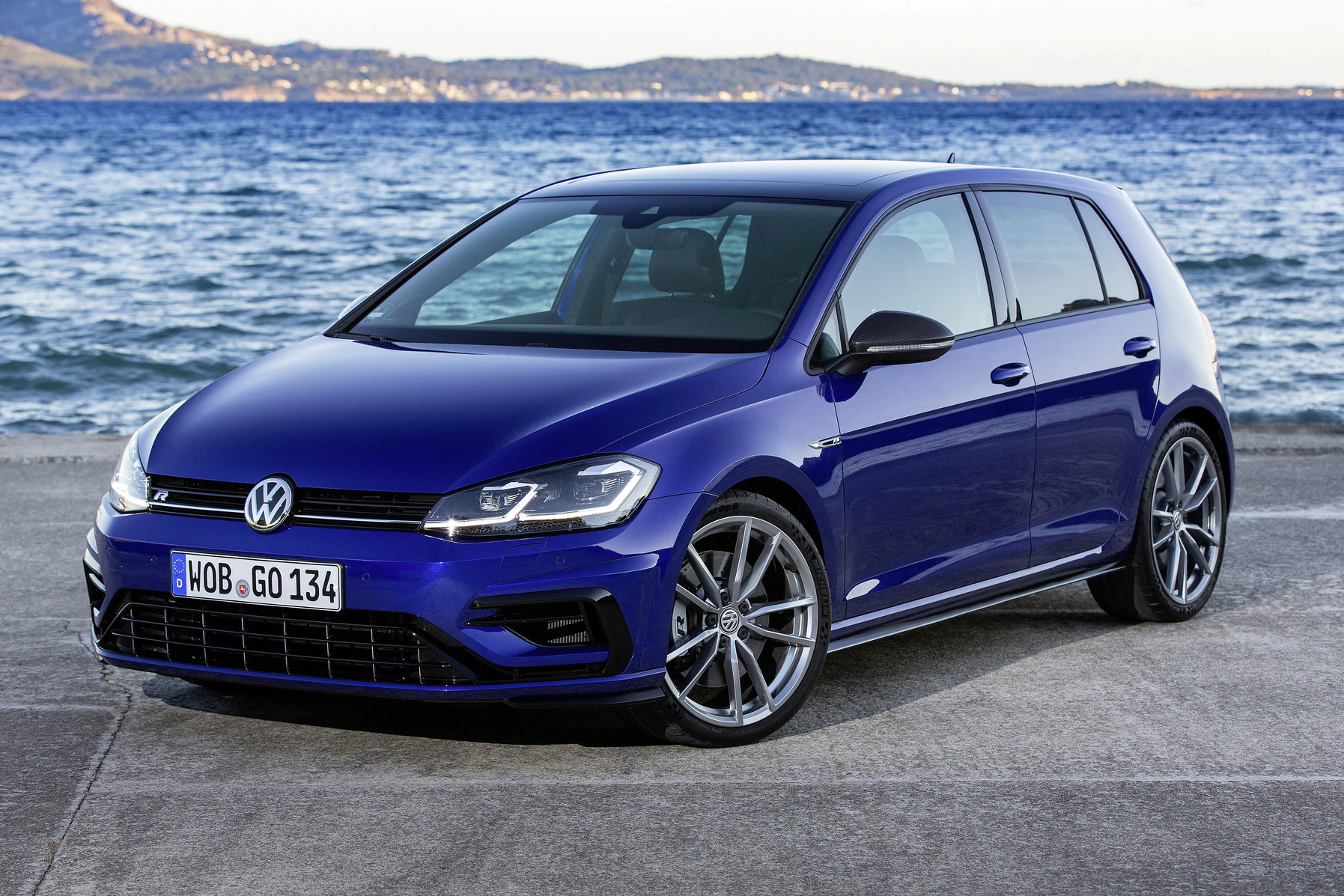 Blauer Volkswagen Golf am Meer, seitlich von vorne fotografiert, mit Bergen im Hintergrund und klarer, sonniger Himmel.