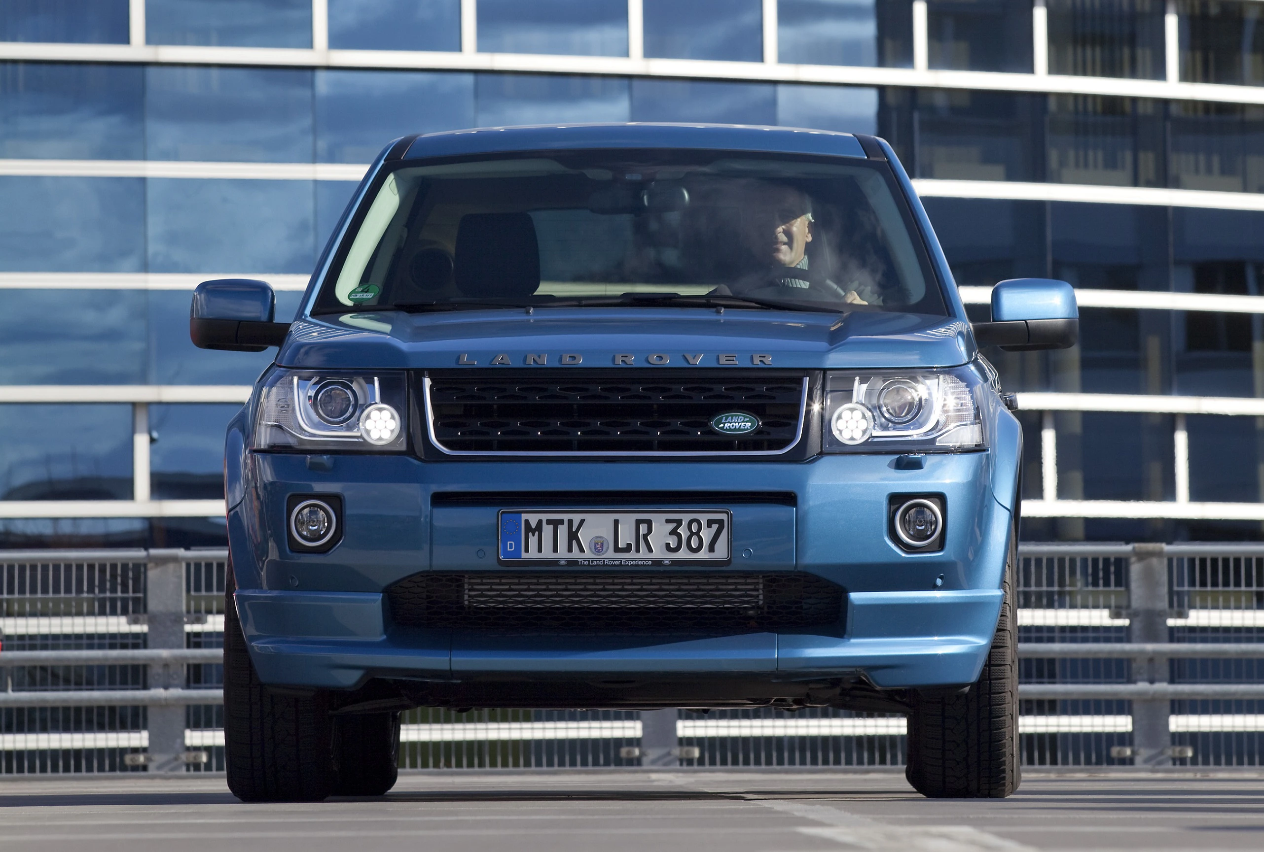 Blauer Land Rover Freelander steht vor einem modernen Gebäude. Nummernschild: MTK LR 387. Mann sitzt am Steuer.