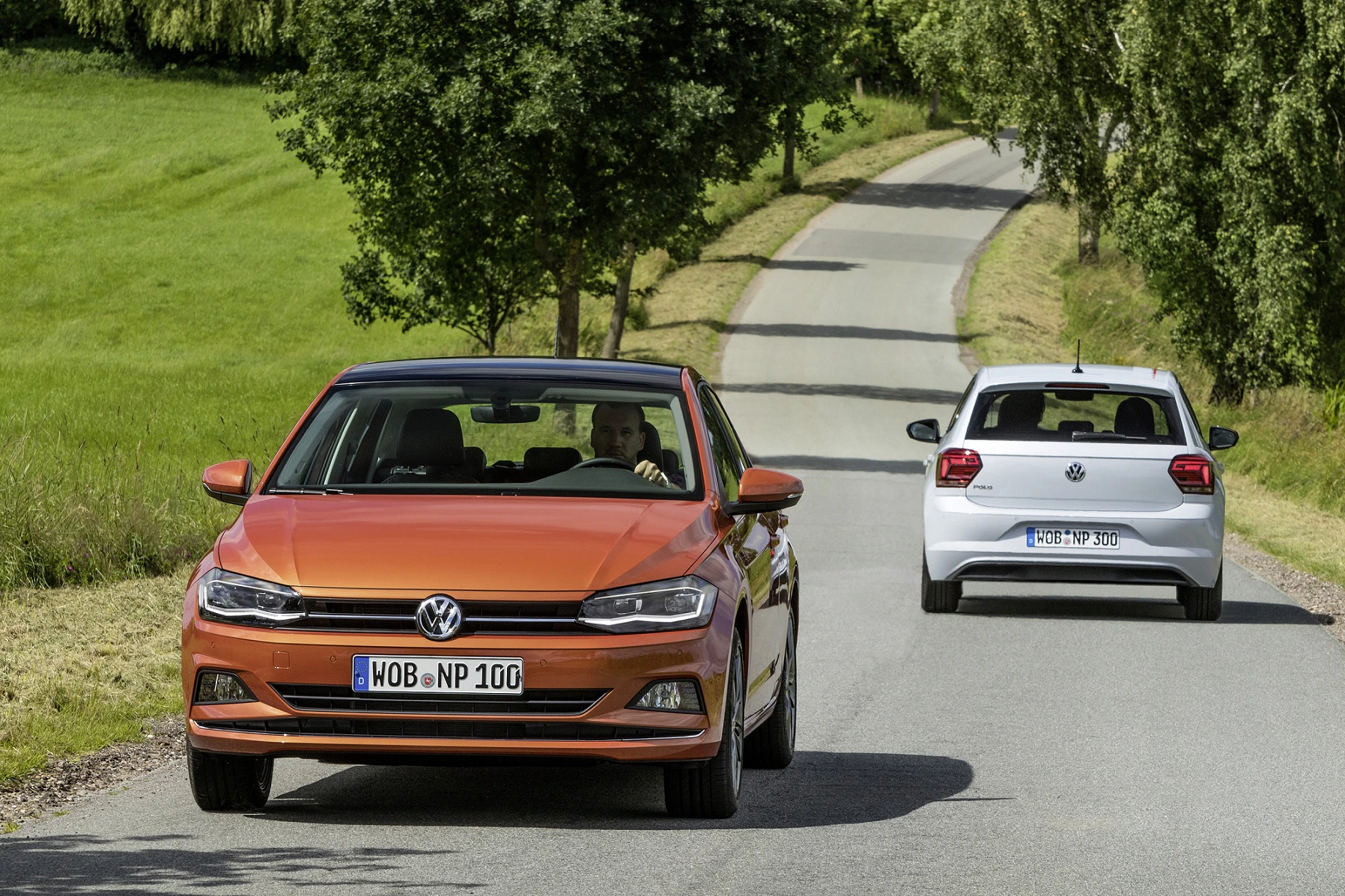 Zwei VW-Autos fahren auf einer ländlichen Straße, umgeben von grüner Landschaft und Bäumen.