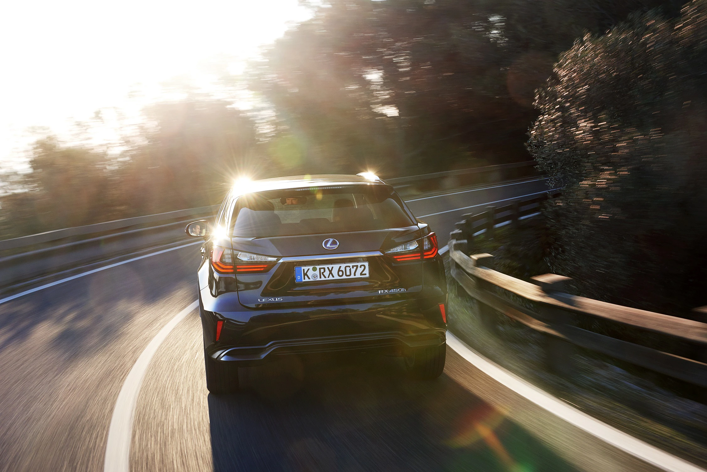 Ein schwarzer Lexus fährt bei Sonnenuntergang auf einer kurvigen Straße durch eine bewaldete Landschaft.