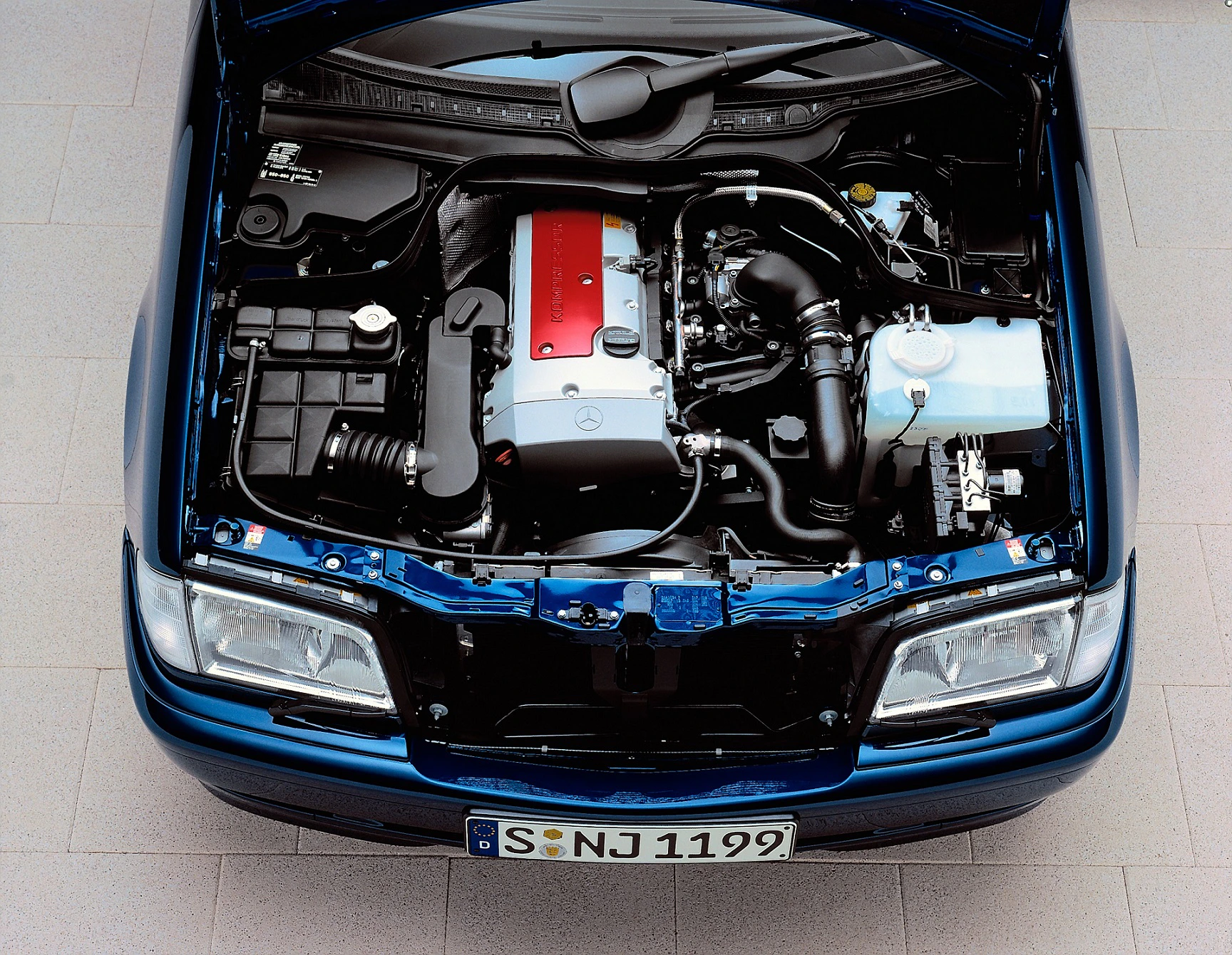 Motorraum eines blauen Autos mit geöffnetem Motorhaube, sichtbar sind Motor, Batterie und diverse Bauteile.