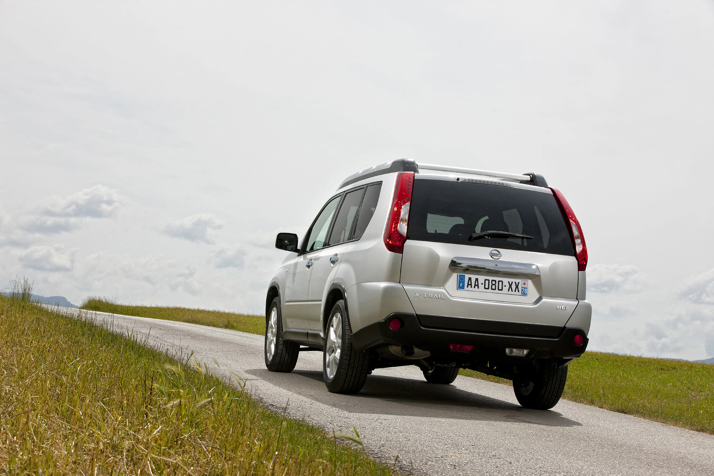 Silberner SUV fährt auf schmaler Landstraße durch grüne Wiesenlandschaft unter bewölktem Himmel.
