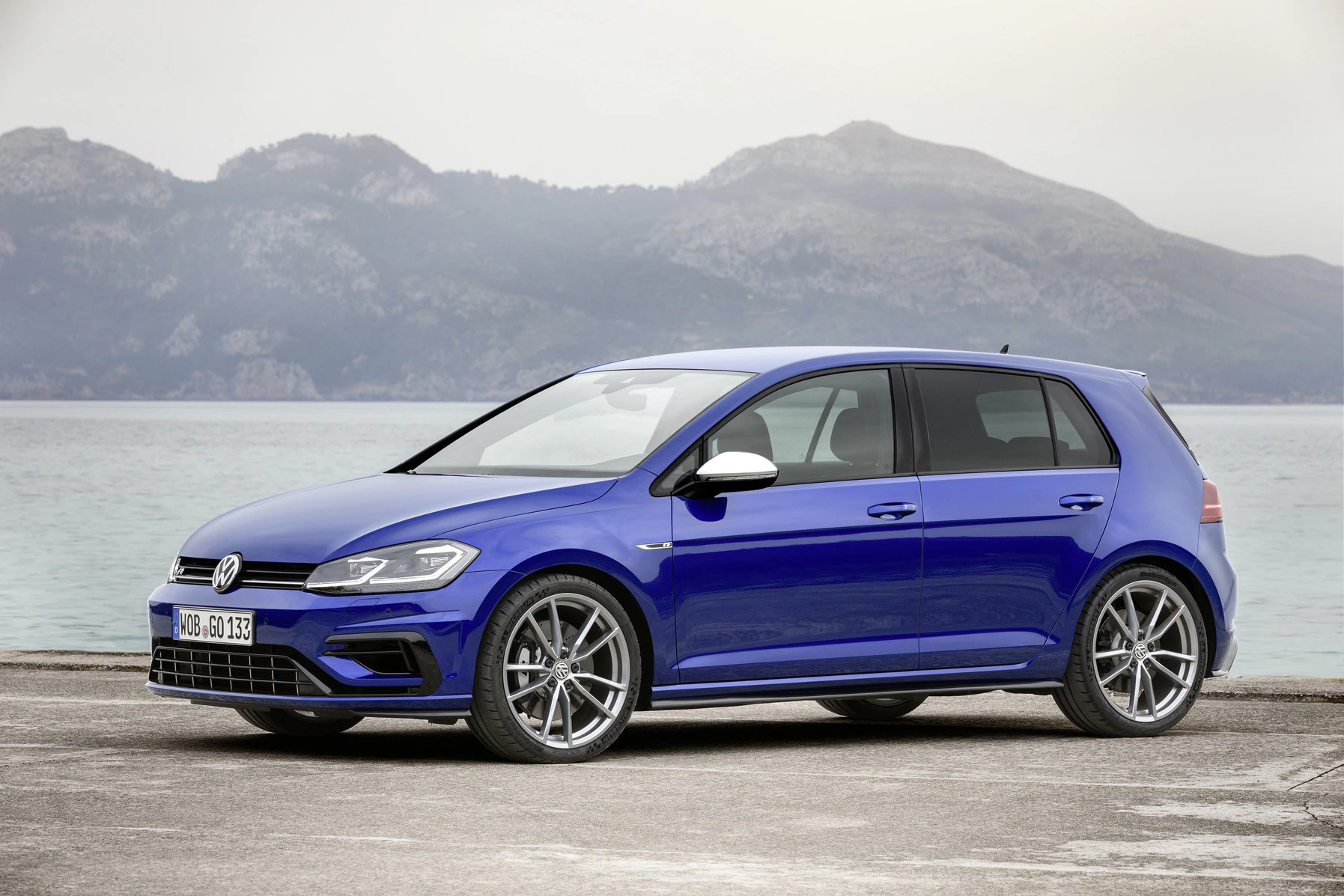 Blauer Volkswagen Golf parkt am Ufer mit Berglandschaft im Hintergrund. Bewölkter Himmel und ruhige Wasseroberfläche.