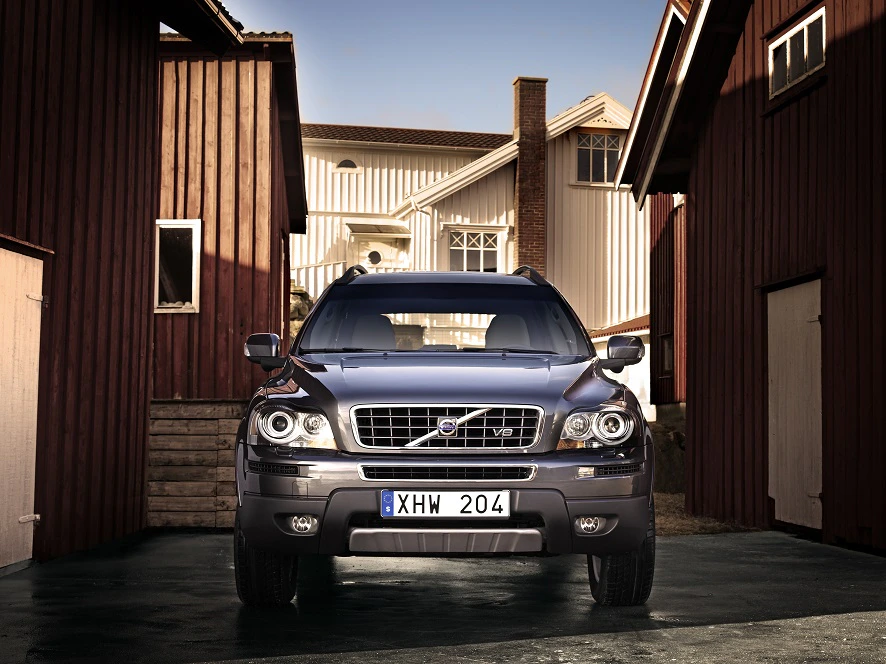 Silberner SUV steht zwischen Holzhäusern. Frontansicht zeigt markantes Design mit Kühlergrill und Scheinwerfern. Blauer Himmel im Hintergrund.
