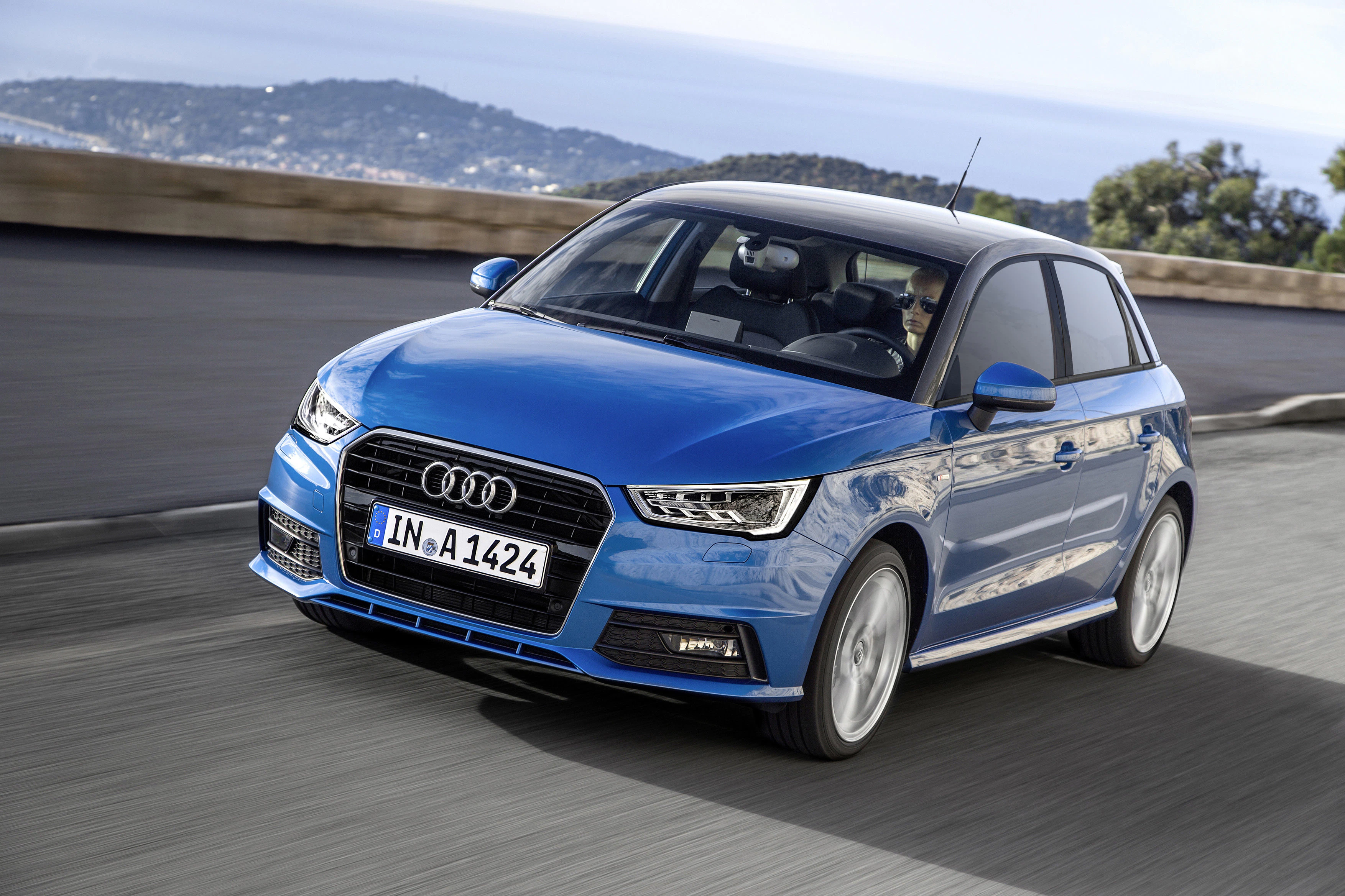 Blauer Audi A1 fährt auf einer Küstenstraße mit Bewuchs im Hintergrund.