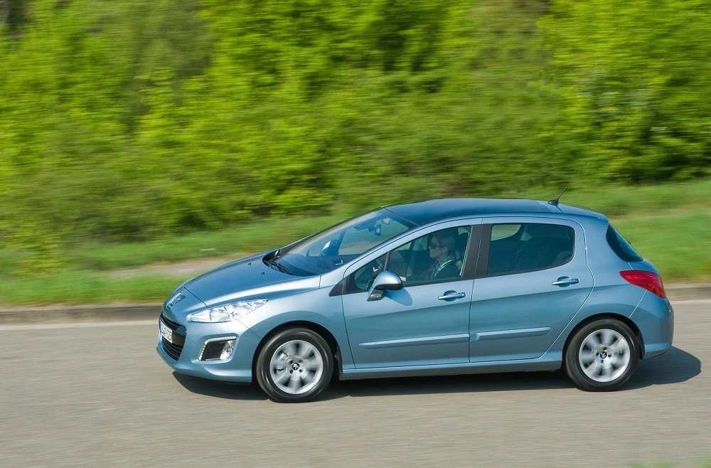 Ein blauer Kleinwagen fährt schnell auf einer Straße, umgeben von grüner Vegetation. Fahrer sichtbar im Innenraum.