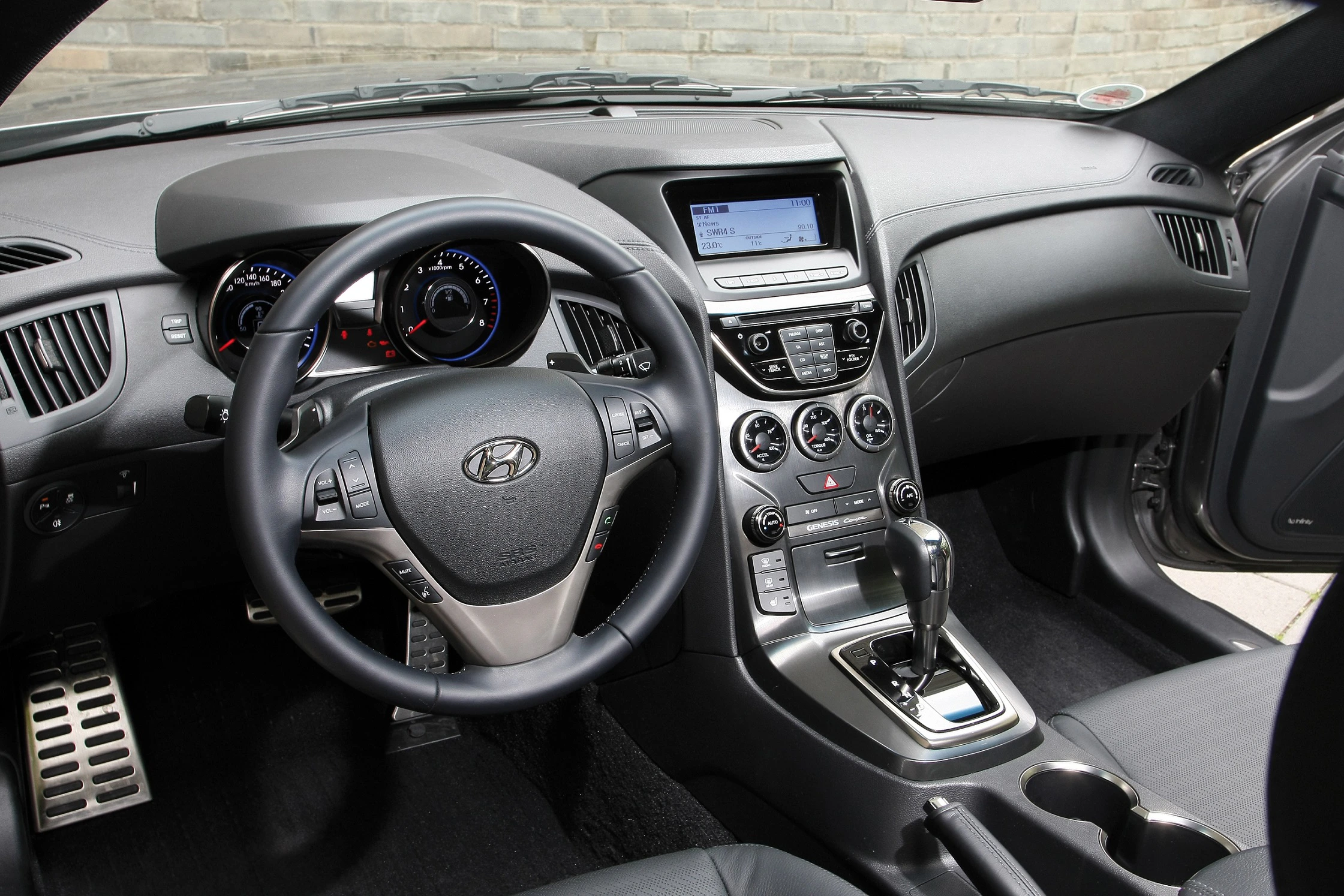 Innenraum eines modernen Autos mit einem Hyundai-Logo auf dem Lenkrad, einem digitalen Display und verschiedenen Bedienelementen auf der Mittelkonsole.