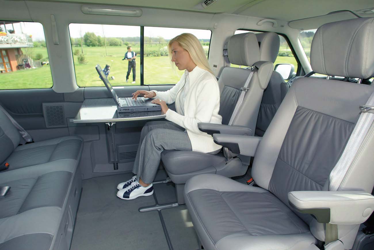 Eine Frau arbeitet im Inneren eines geräumigen Autos an einem Laptop. Die Sitze sind grau und der Hintergrund zeigt eine Grünfläche.