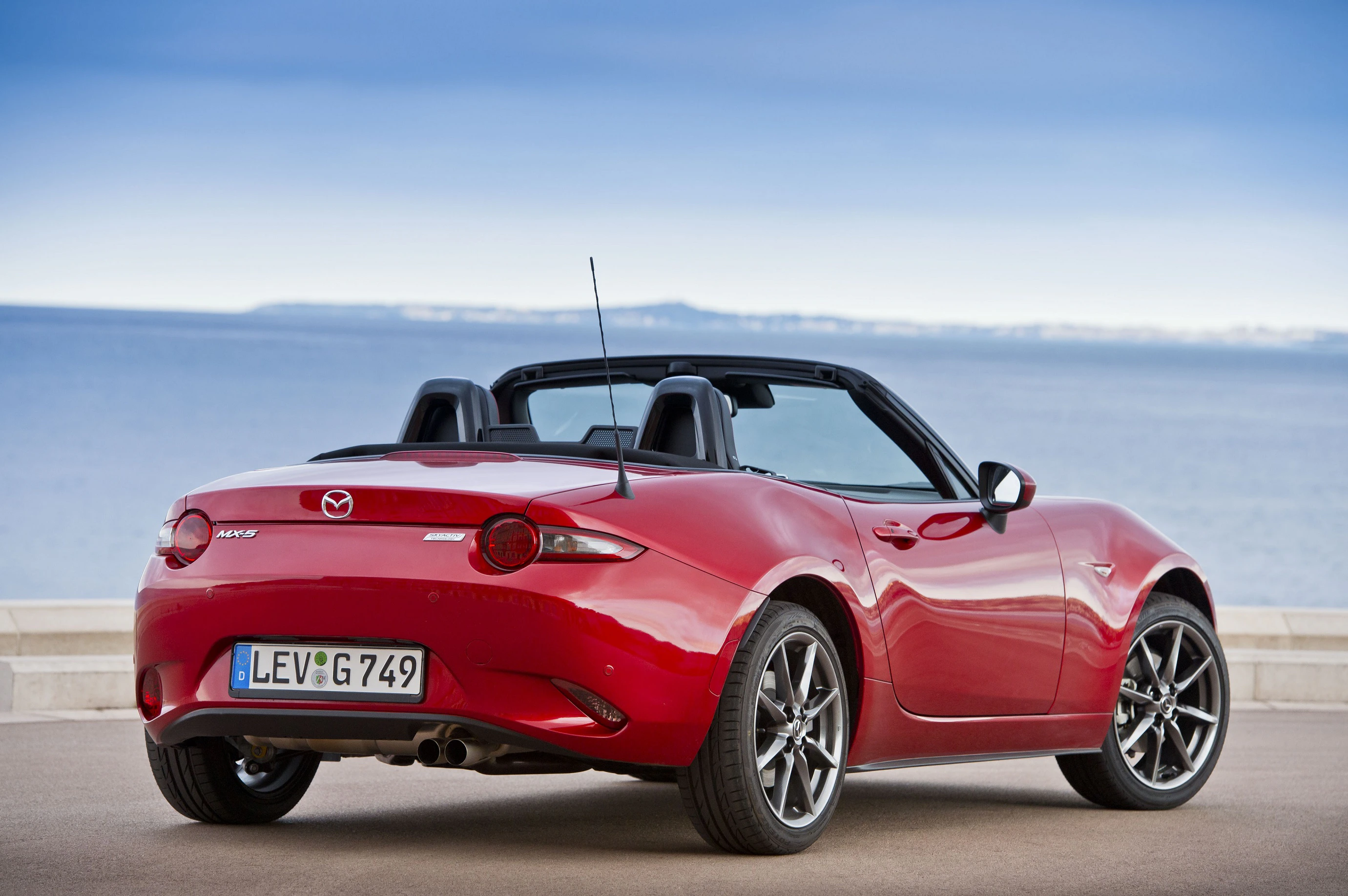 Roter Mazda MX-5 Cabrio, geparkt vor einer Küstenkulisse mit blauem Himmel.