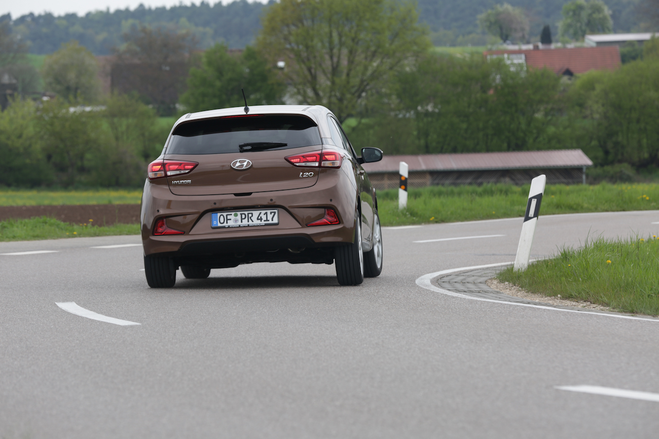 Ein brauner Hyundai i20 fährt auf einer kurvigen Landstraße; im Hintergrund Grünflächen und bewaldete Hügel.