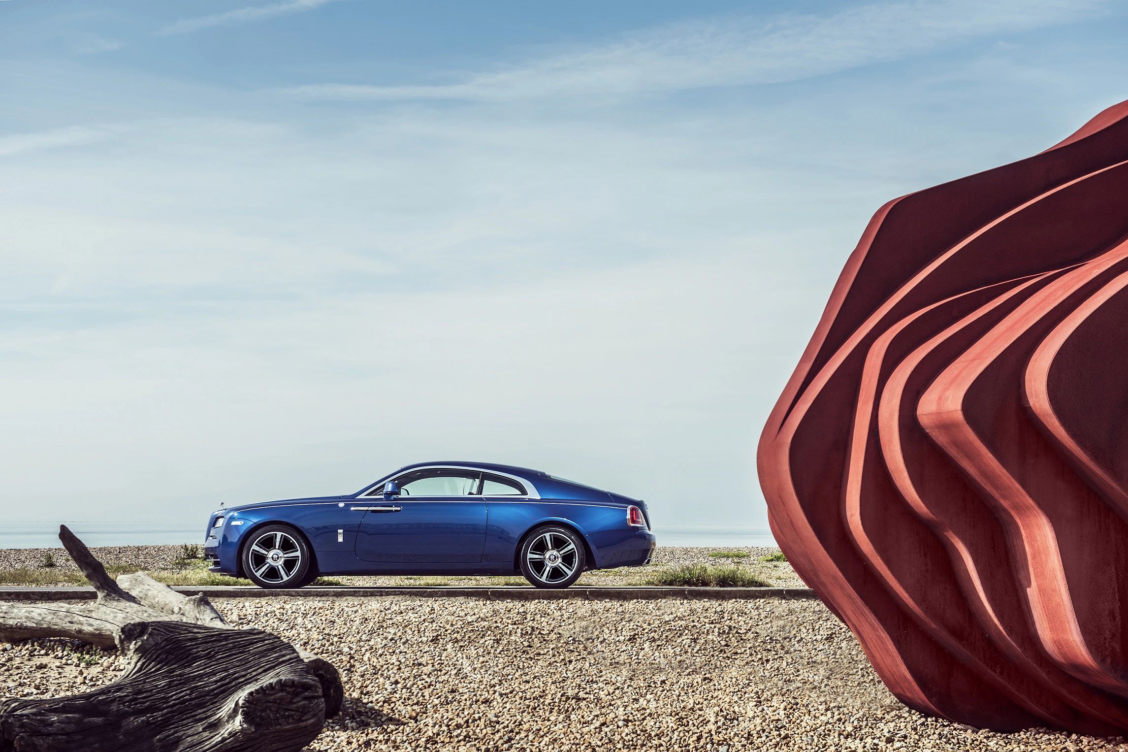 Blaues Auto neben großer roter Skulptur an einem Küstenstreifen mit Kiesboden und Holzelementen, unter klarem Himmel.