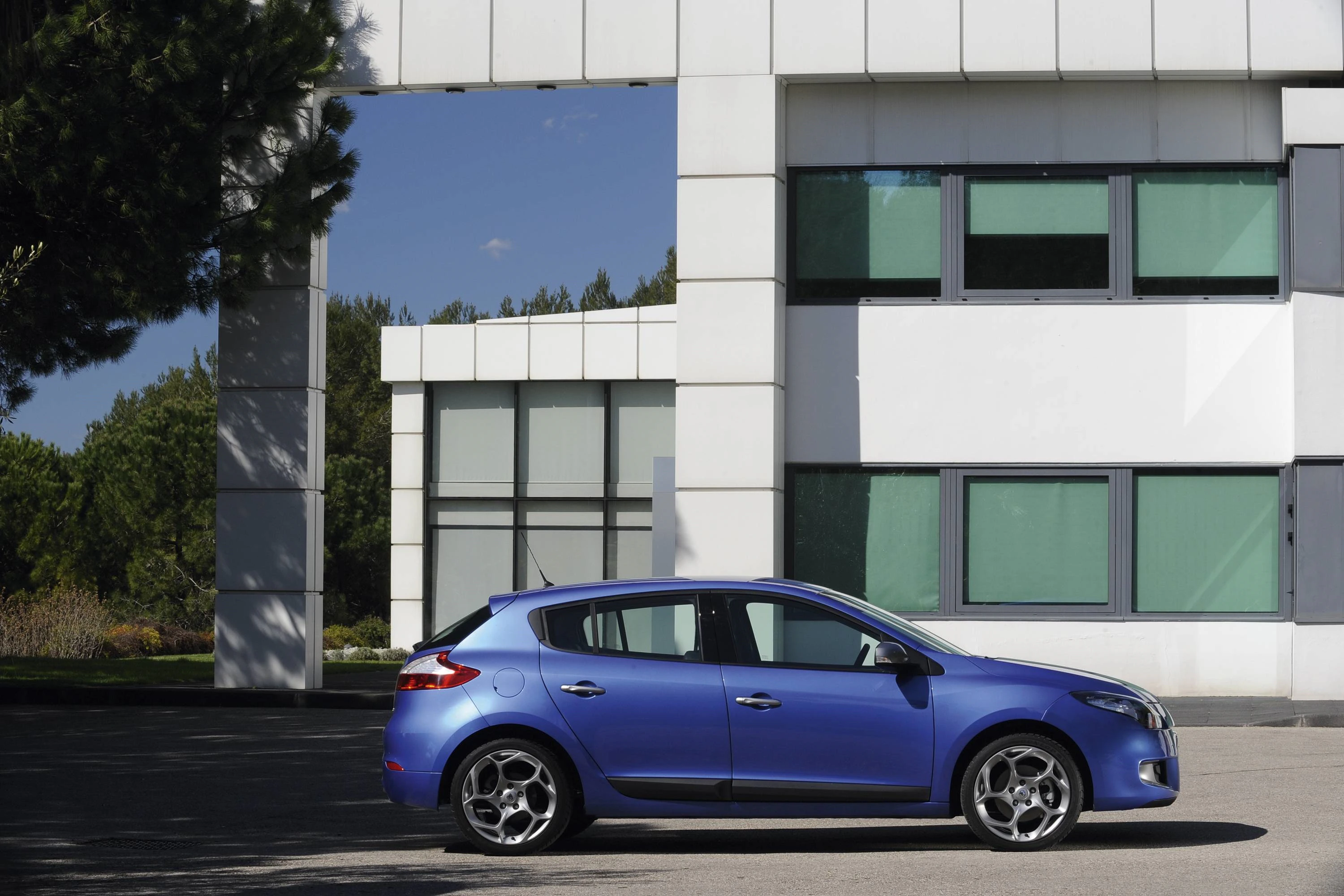 Blaues Auto vor modernem, weißem Gebäude mit großen Fenstern, umgeben von Bäumen und klarem Himmel.