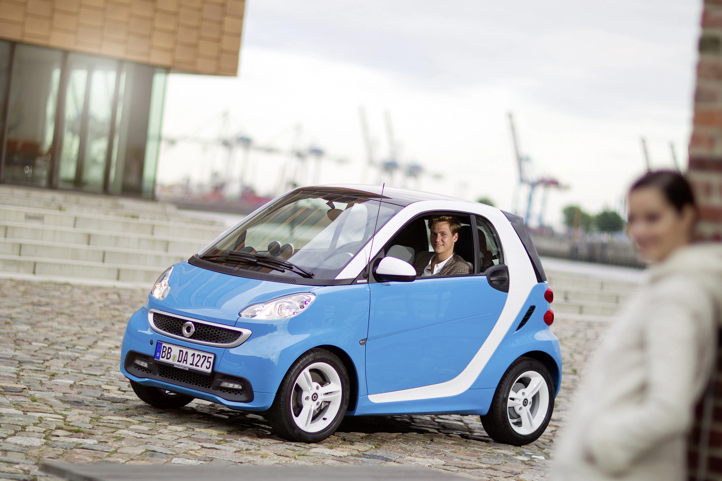 Ein Mann sitzt in einem blauen Smart Fortwo auf einem gepflasterten Platz. Im Hintergrund stehen Hafenkräne, eine Frau ist im Vordergrund unscharf.