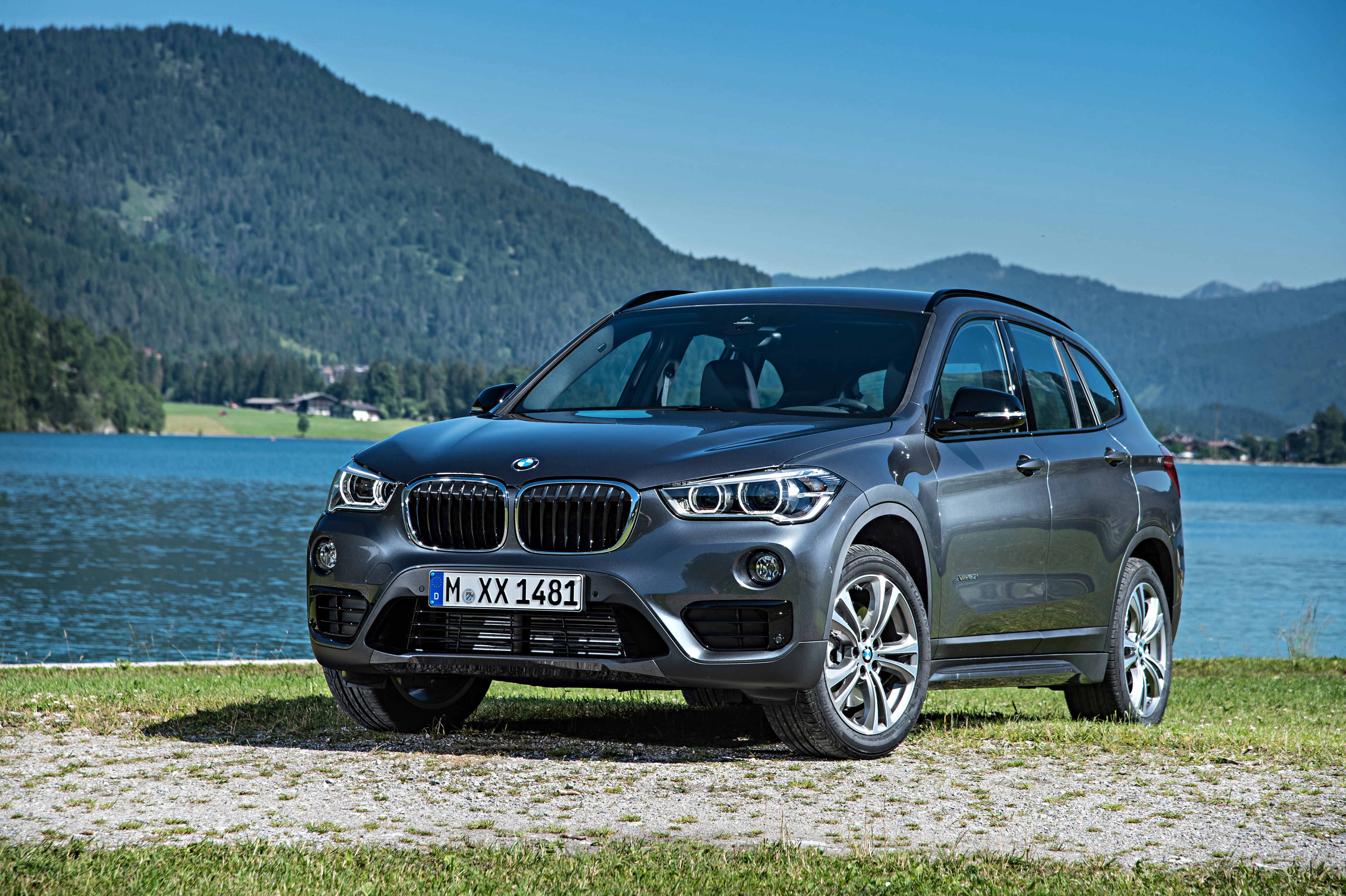 Silbergrauer BMW SUV steht vor einem ruhigen See mit bewaldeten Bergen im Hintergrund unter klarem, blauem Himmel.
